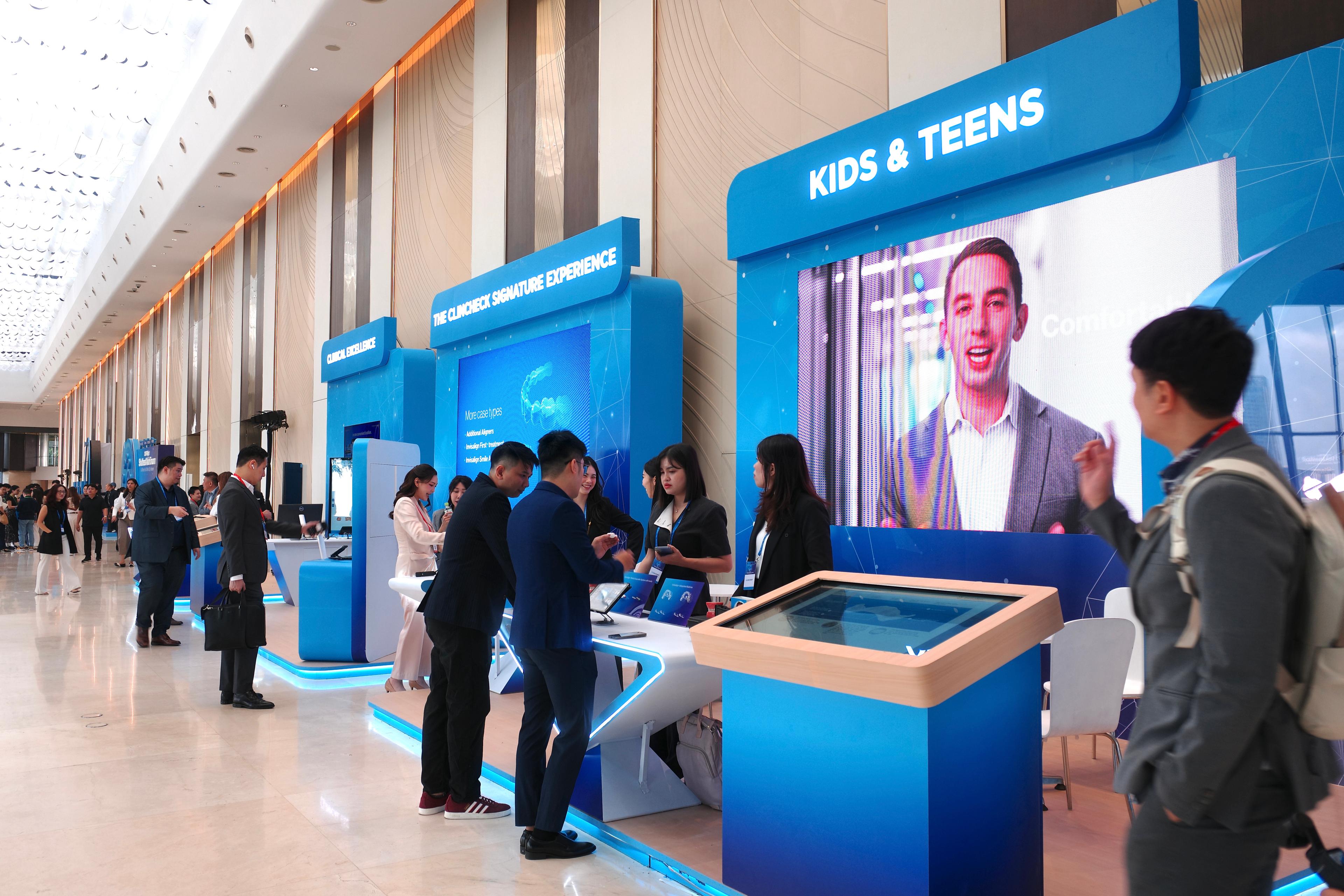 Khu trưng bày của diễn đàn với các gian mang bảng “CLINICAL EXCELLENCE”, “THE CLINCHECK SIGNATURE EXPERIENCE” và “KIDS & TEENS”; đại biểu đứng thử thiết bị và máy tính bảng tại từng gian.