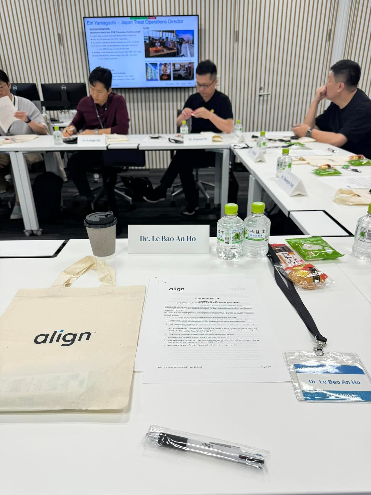 Bàn hội thảo tại văn phòng Align Technology với biển tên “Dr. Le Bao An Ho”, thẻ dự chương trình và tài liệu đặt trước chỗ ngồi; màn hình phía sau đang trình chiếu phần báo cáo của Japan Treat Operations Director.