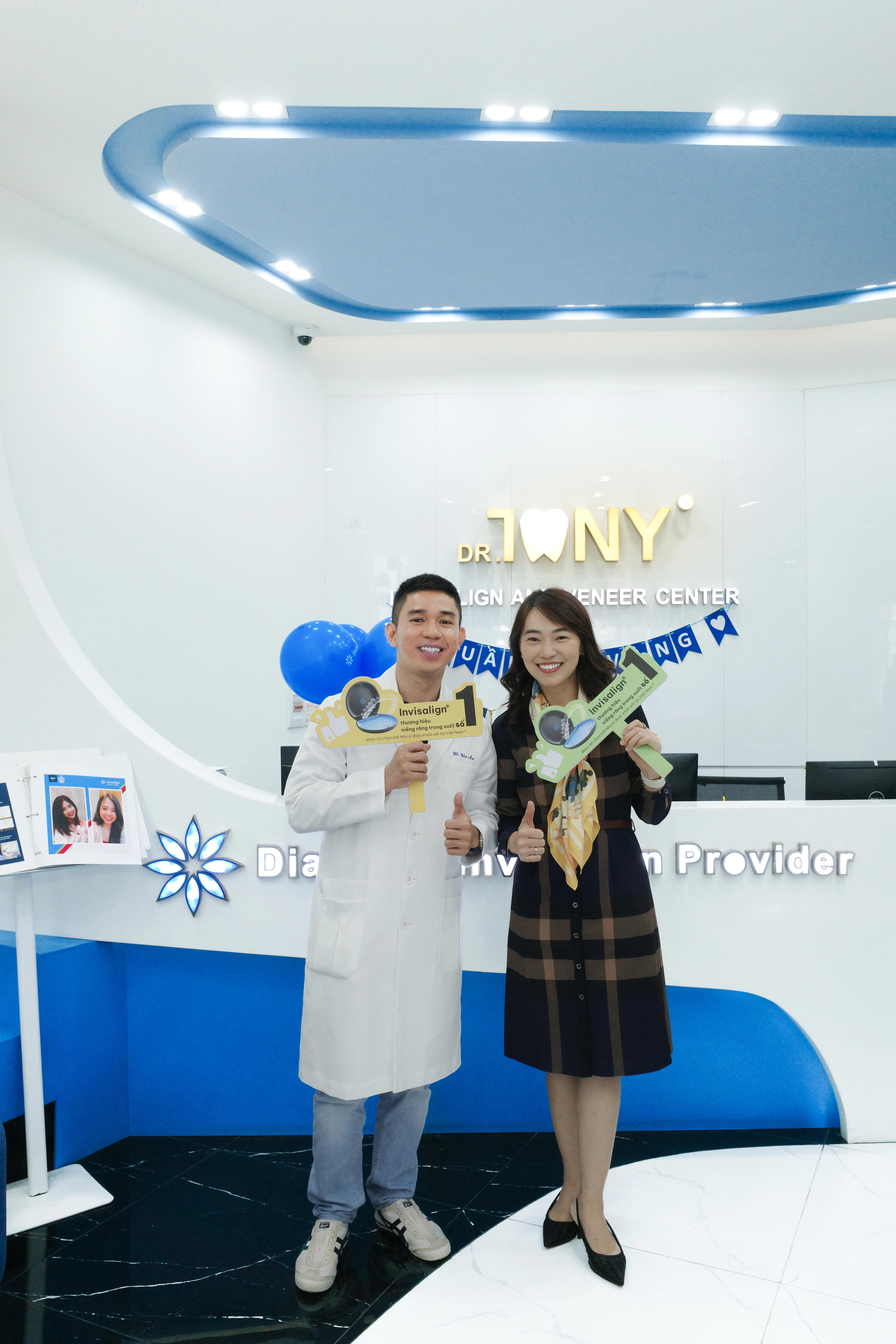 Một bác sĩ mặc áo blouse trắng và một khách đứng ở sảnh phòng khám, mỗi người cầm một bảng cổ vũ in chữ “Invisalign”; phía sau là bảng hiệu vàng “DR. TONY — INVISALIGN AND VENEER CENTER”, bóng bay xanh và dây cờ trang trí.