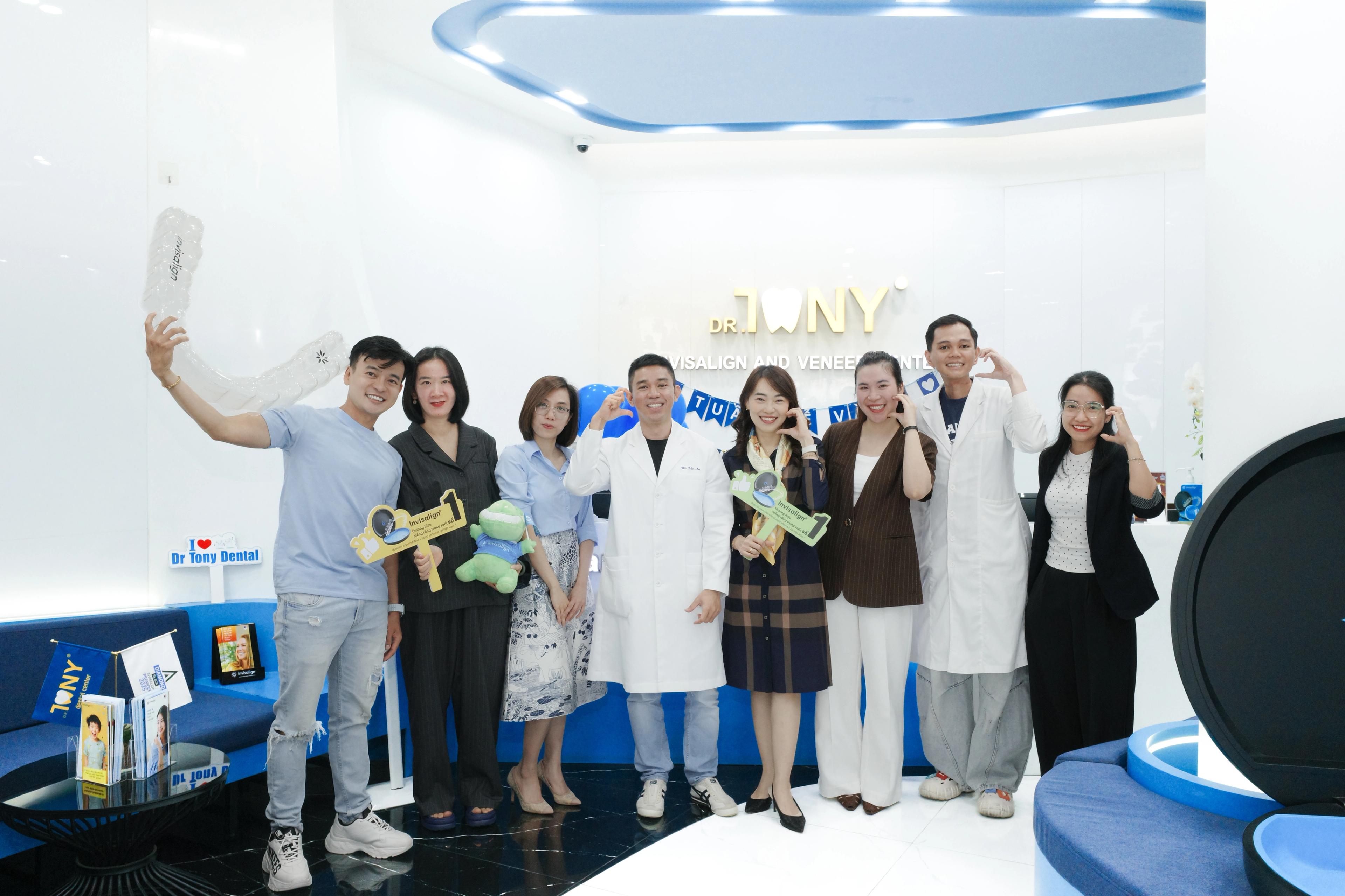 Chín người đứng thành hàng ở sảnh phòng khám: người ngoài cùng bên trái giơ cao mô hình khay trong suốt, hai người cầm bảng cổ vũ “Invisalign”, một người cầm thú bông hình răng; phía sau là bảng hiệu vàng “DR. TONY — INVISALIGN AND VENEER CENTER” và dây cờ trang trí.