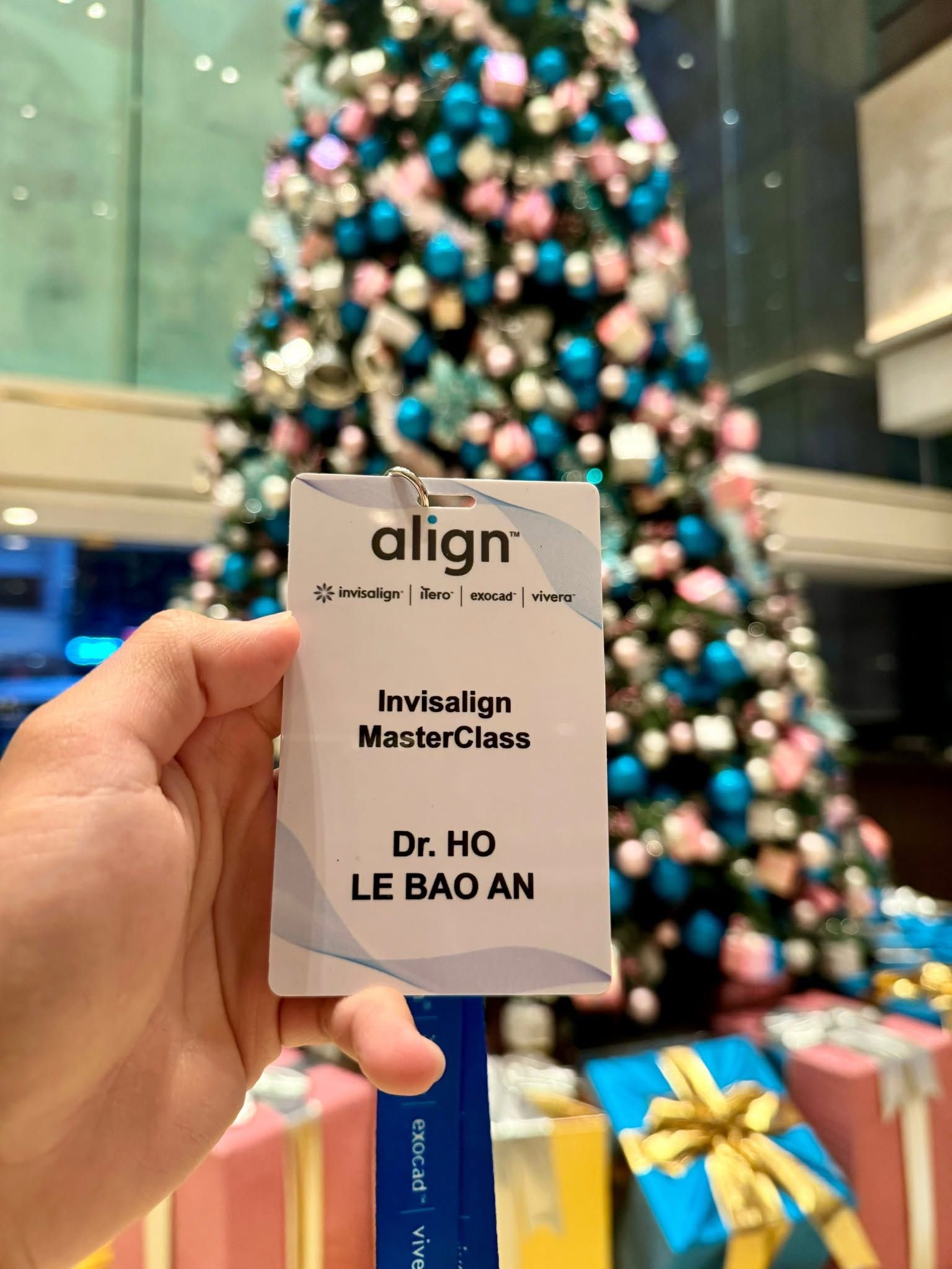 Thẻ tên dự chương trình ghi “Invisalign MasterClass – Dr. Ho Le Bao An” cầm trên tay, phía sau là sảnh khách sạn nơi diễn ra khóa học.