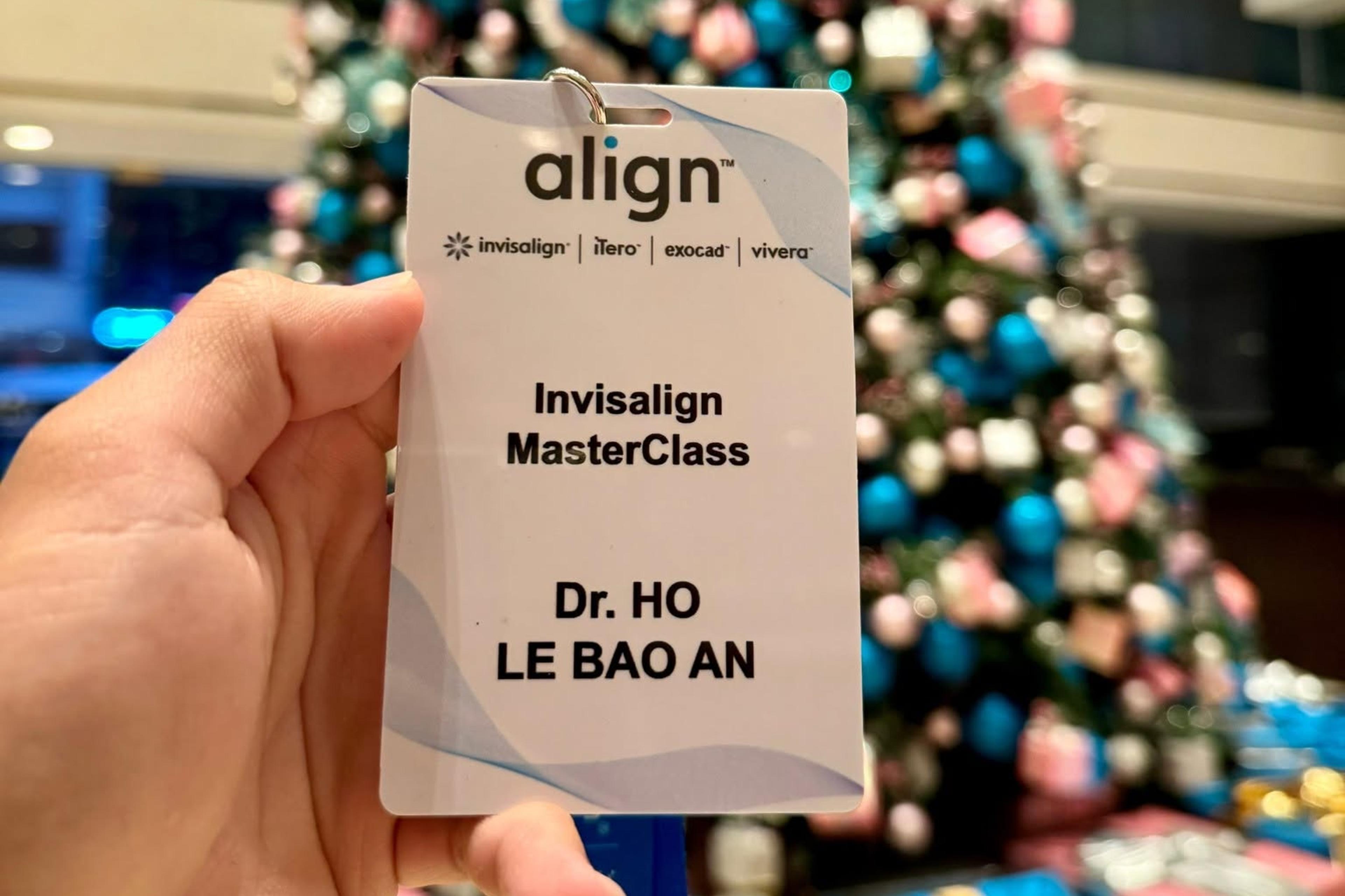Thẻ tên dự chương trình ghi “Invisalign MasterClass – Dr. Ho Le Bao An” cầm trên tay, phía sau là sảnh khách sạn nơi diễn ra khóa học.