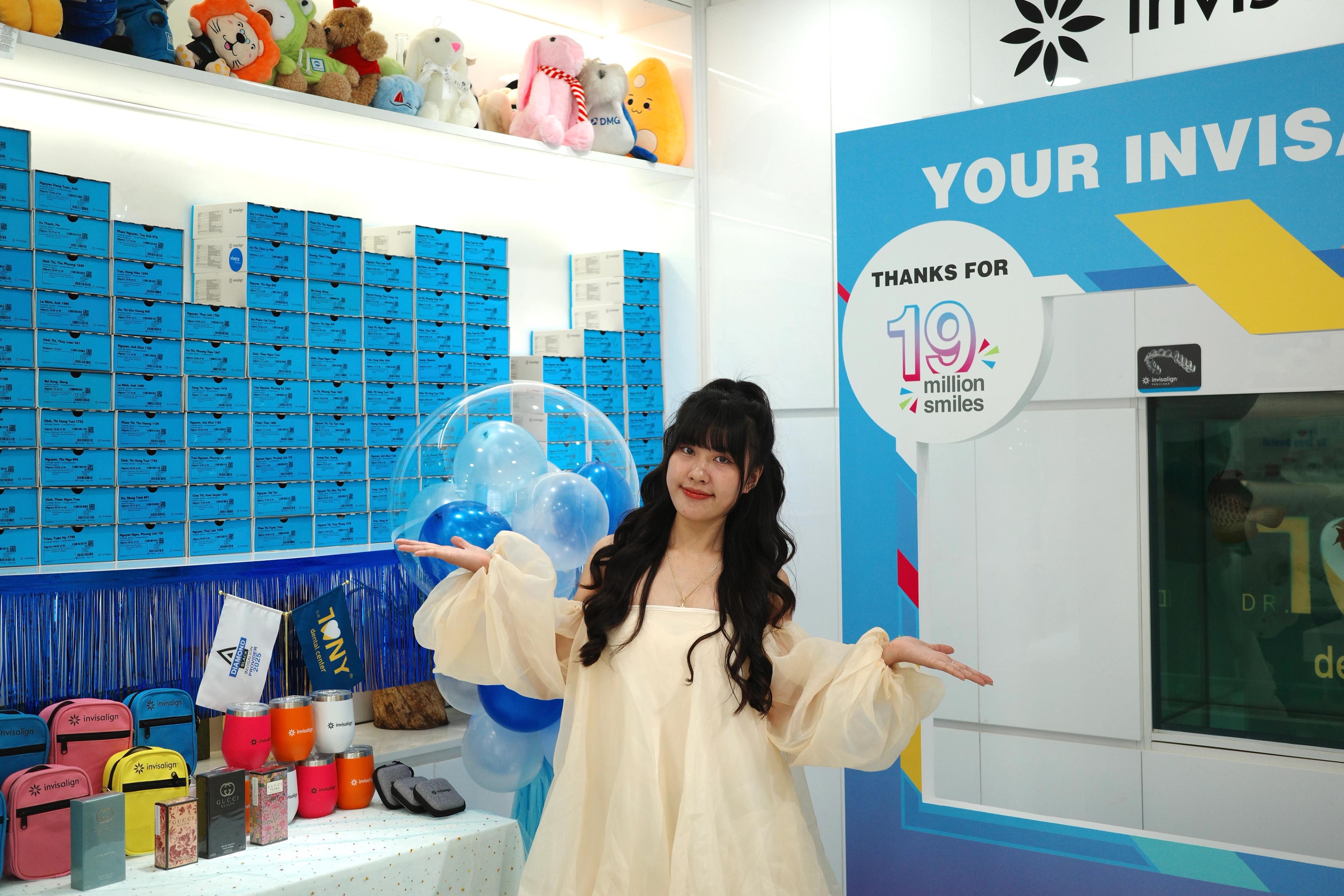 Một khách mời đứng dang tay giữa bàn quà và mảng tường xanh in “THANKS FOR 19 million smiles”; phía trên bức tường hộp Invisalign là kệ thú bông.