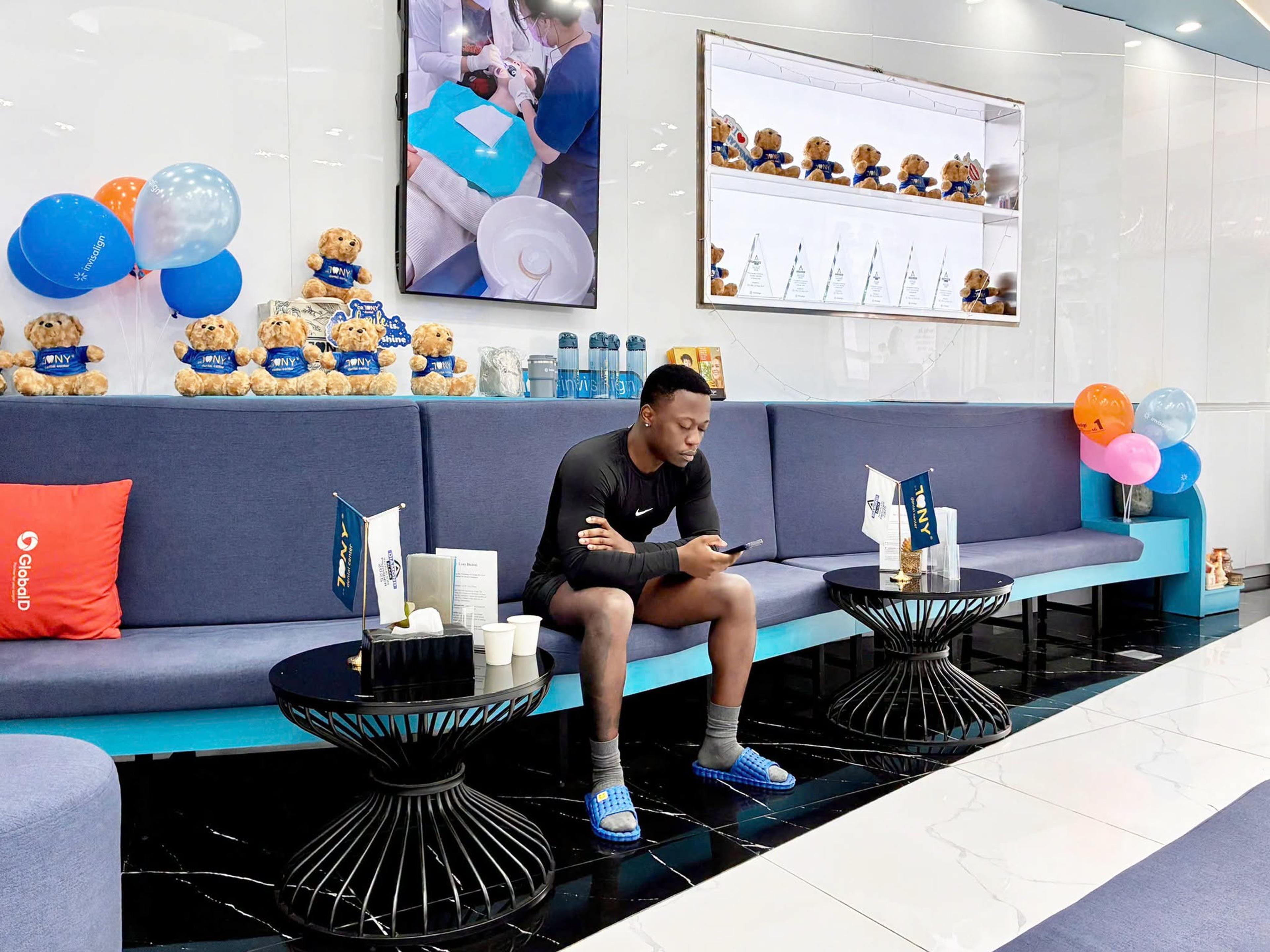 Một khách ngồi chờ trên ghế sofa xanh, phía trên là hàng gấu bông mặc áo in logo Dr. Tony và chùm bóng bay; trên tường là màn hình chiếu cảnh điều trị và tủ kính trưng bày các kỷ niệm chương pha lê.