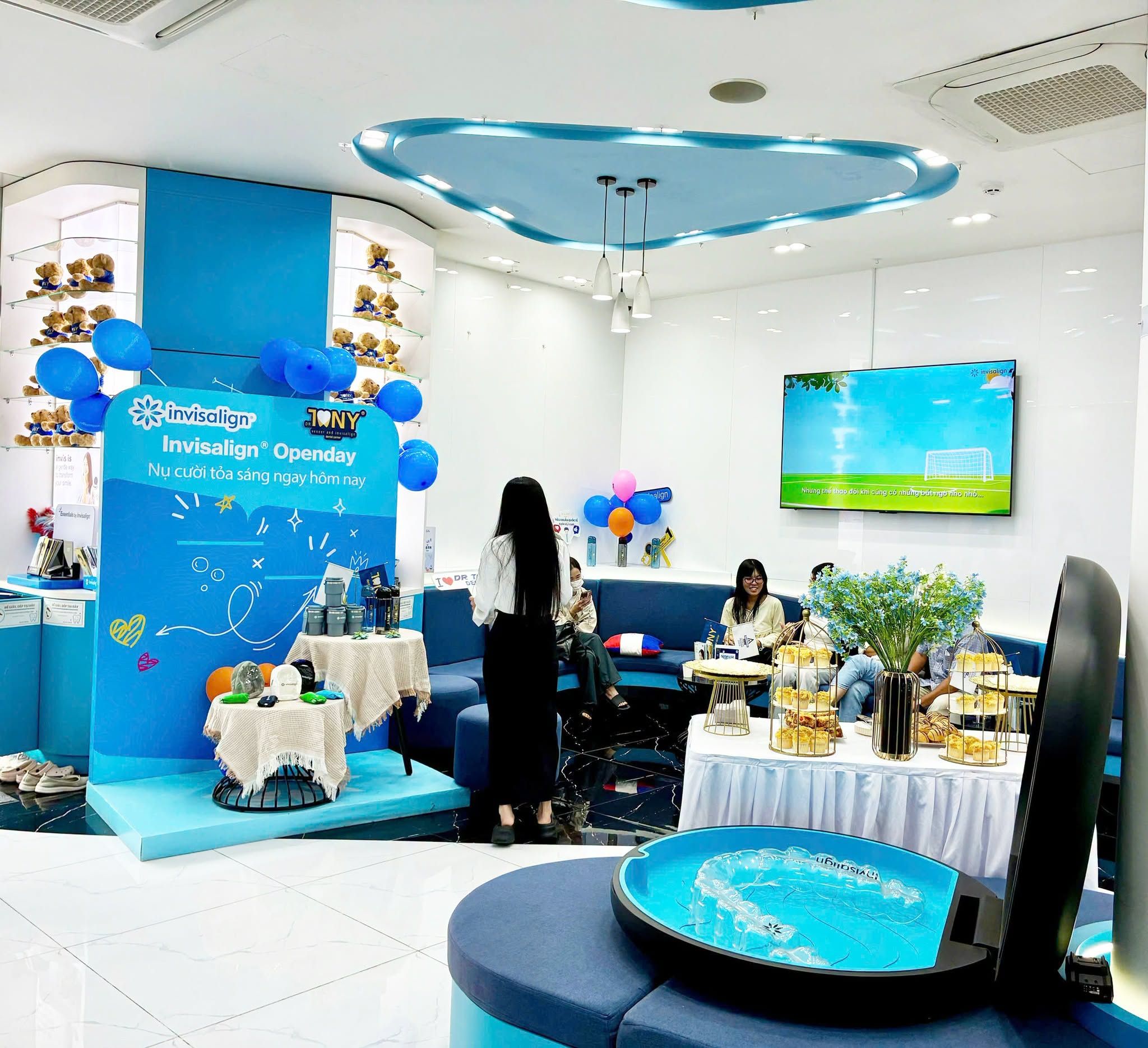Góc check-in "Invisalign® Openday — Nụ cười tỏa sáng ngay hôm nay" mang logo Invisalign và Dr. Tony, cạnh bàn quà bày bình giữ nhiệt; phía sau là khu ghế chờ và kệ gấu bông, phía trước là mô hình hộp đựng khay Invisalign cỡ lớn đang mở nắp.