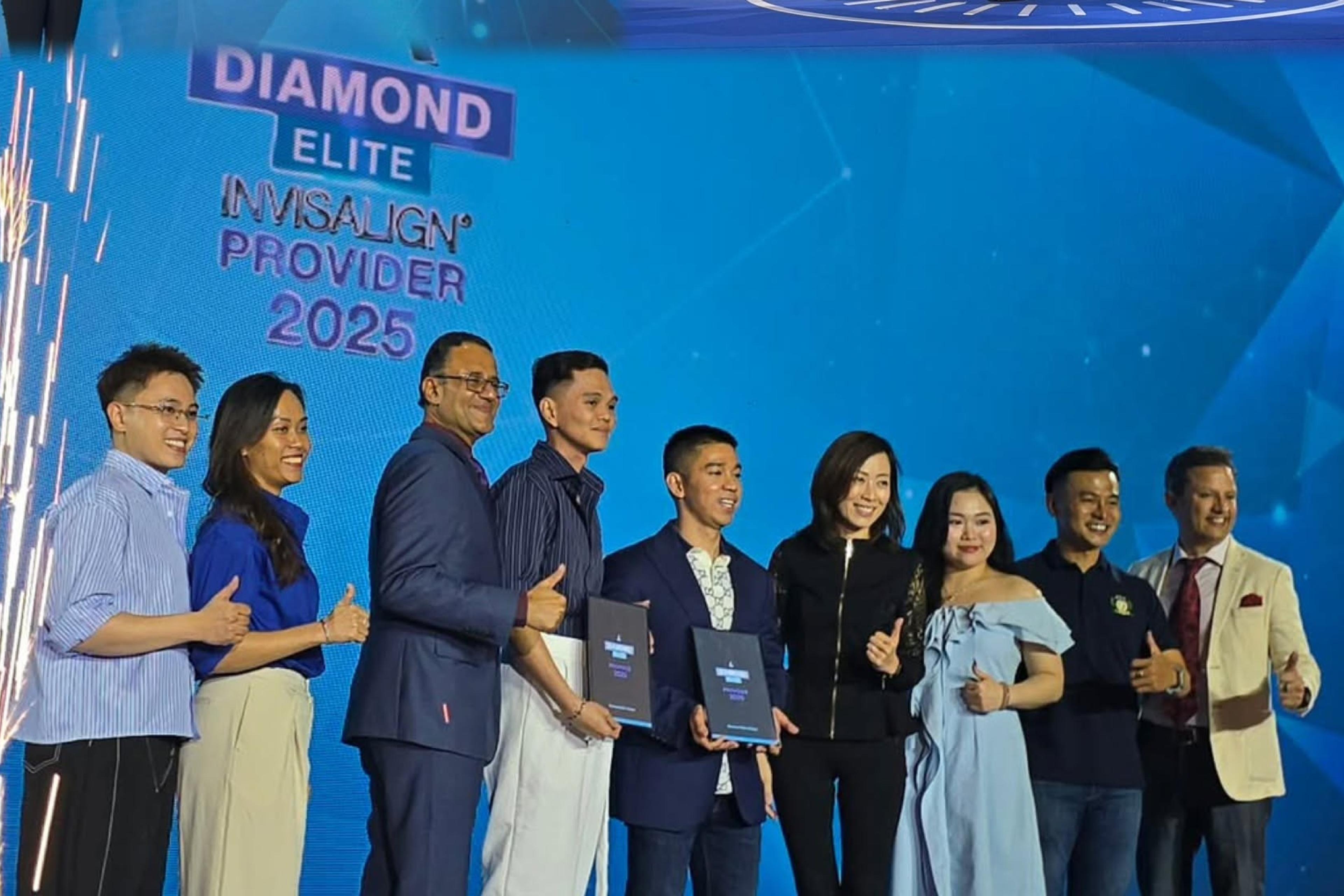 Chín người đứng thành hàng trên sân khấu, một dải pháo sáng ở mép trái, hai người ở giữa cầm bảng chứng nhận; màn hình phía sau ghi “Diamond Elite Invisalign Provider 2025”.