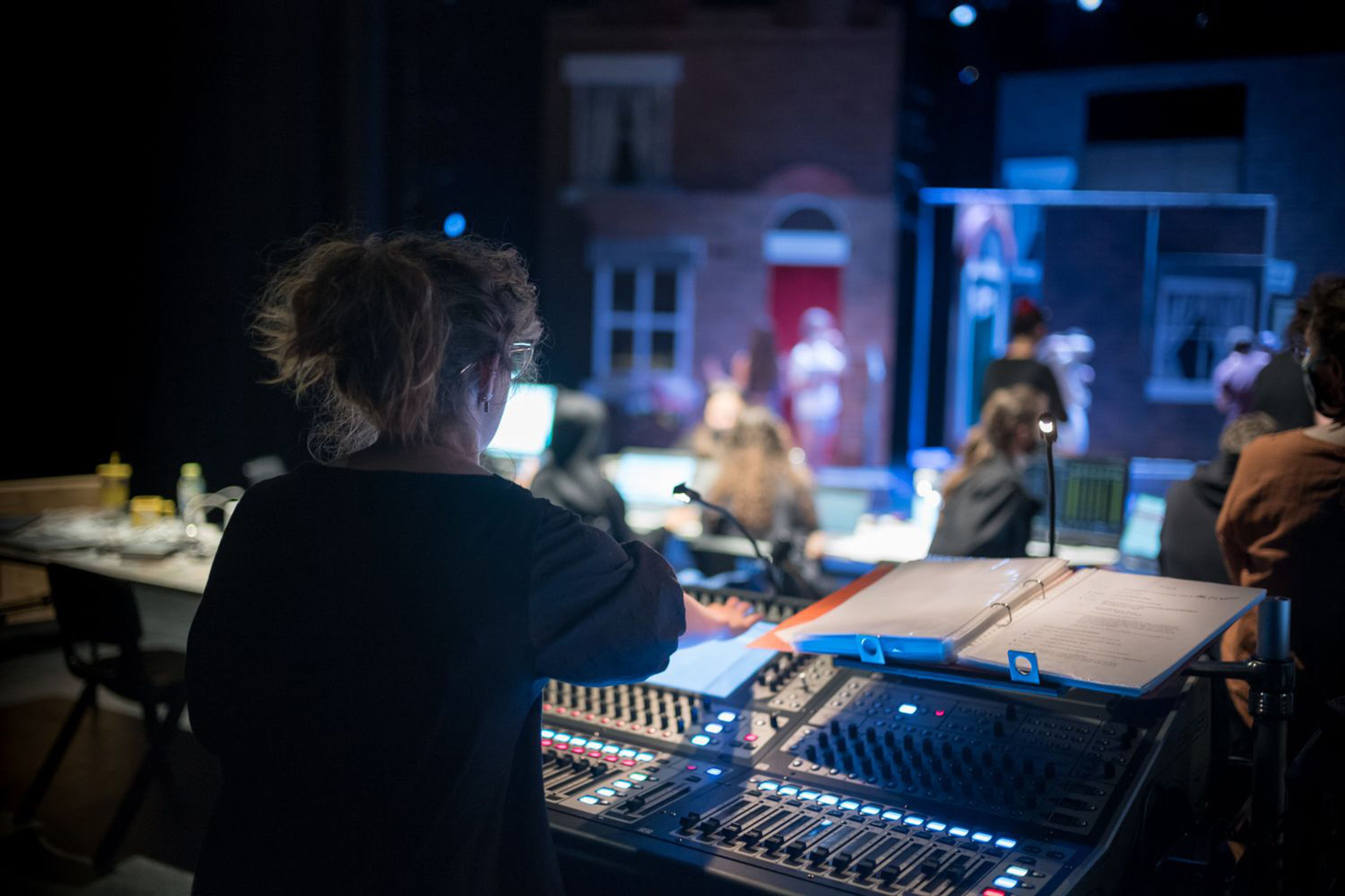 A person with curly hair seen from behind, working at a sound desk in a theatre . In the background, a stage is set with a house facade and people in rehearsal.