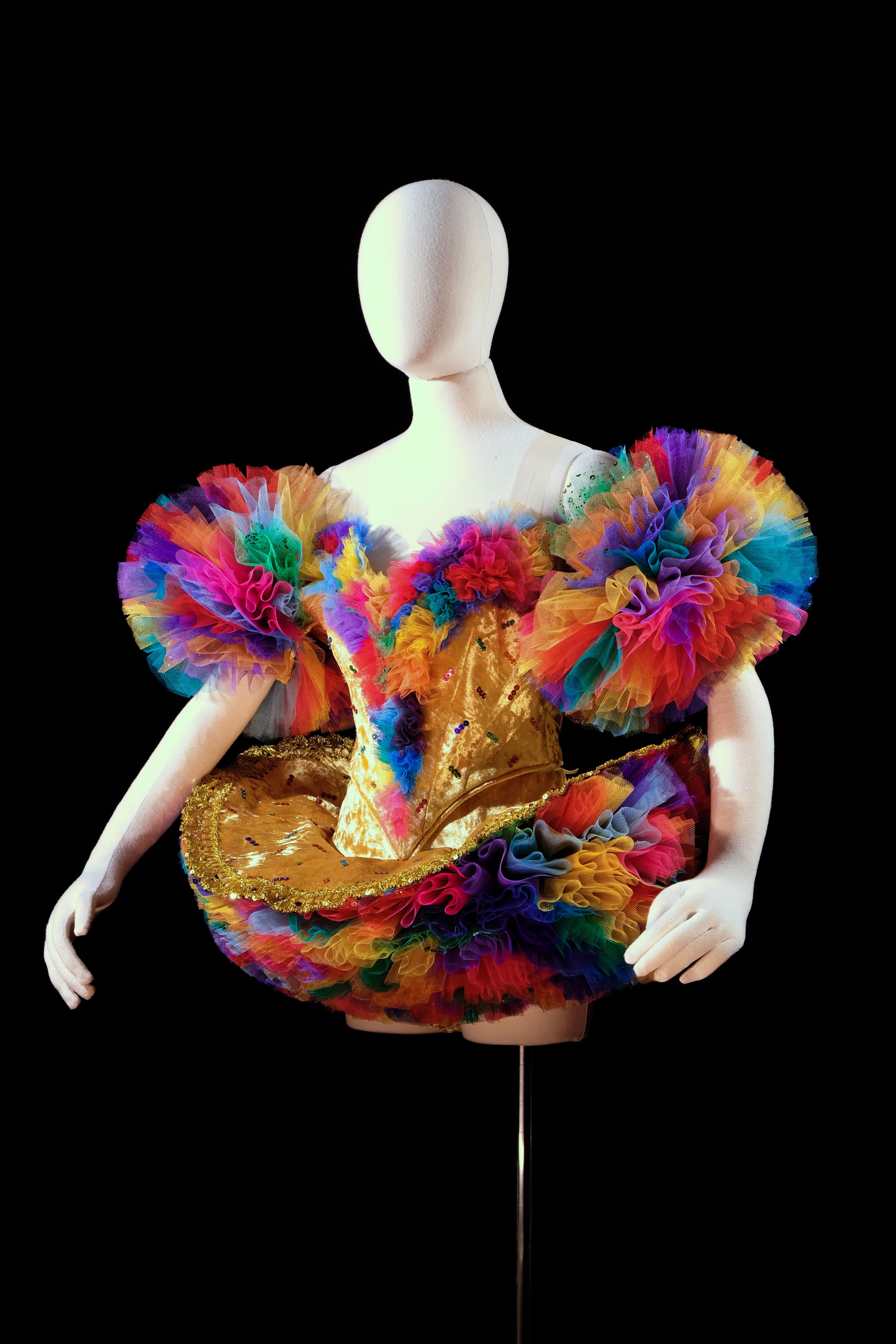 A mannequin dressed in an elaborate costume featuring a golden bodice adorned with sequins and vibrant, multicolored tulle ruffles. The outfit includes dramatic puffed sleeves and a flared skirt