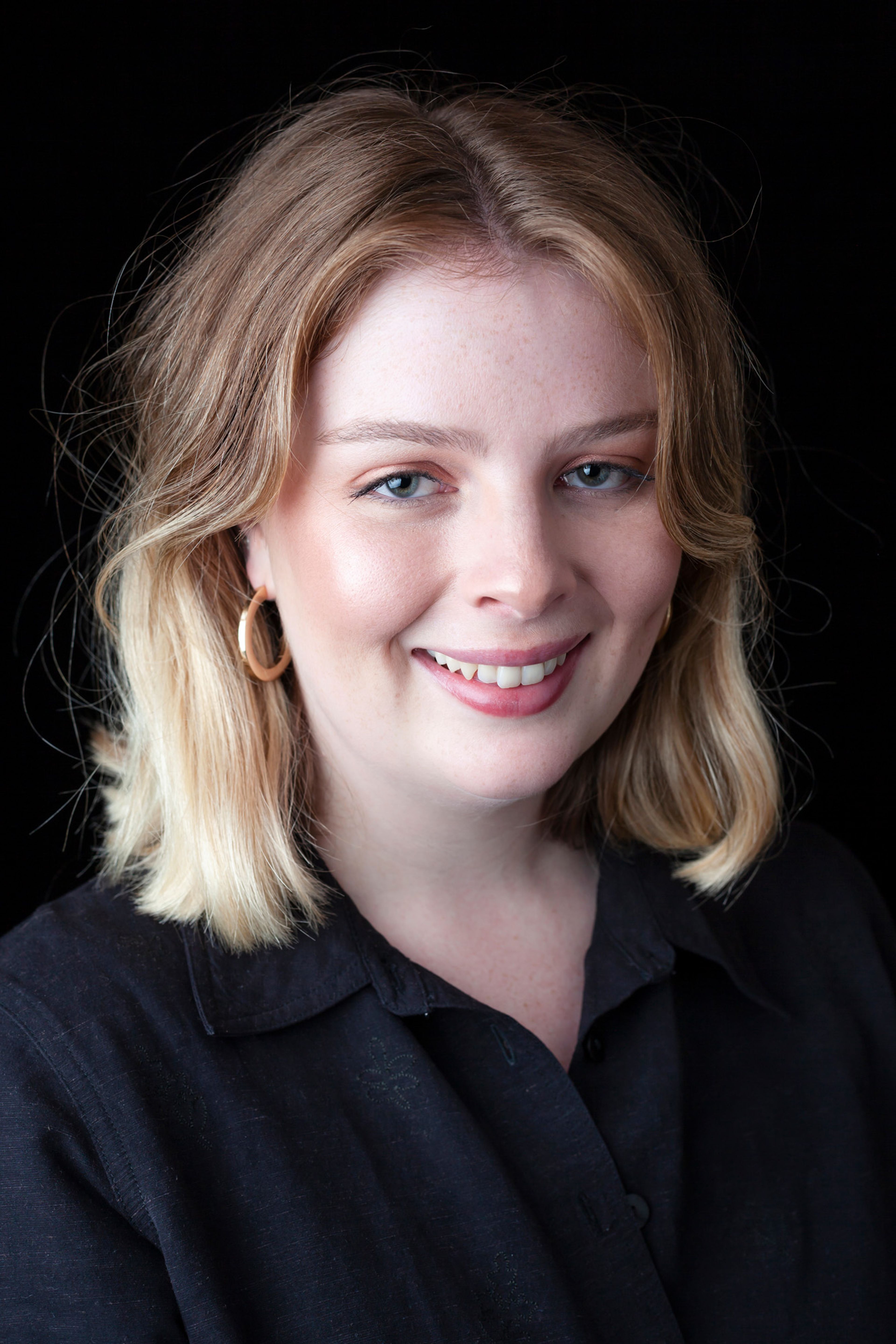 Portrait of a woman with shoulder-length blonde hair, wearing a black shirt and gold hoop earring