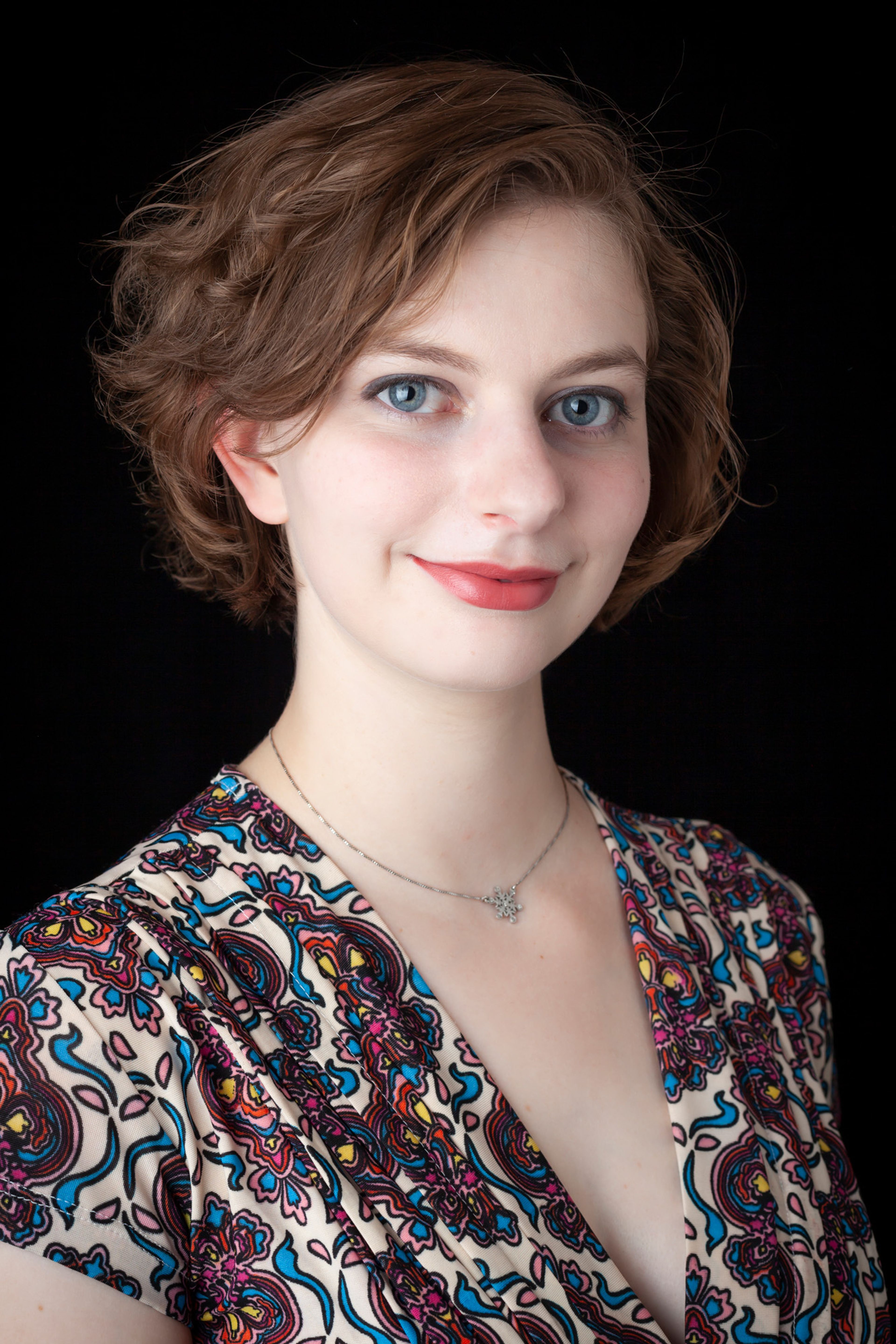 Portrait of a person with short, wavy brown hair, wearing a colorful patterned top and a silver necklace, smiling softly