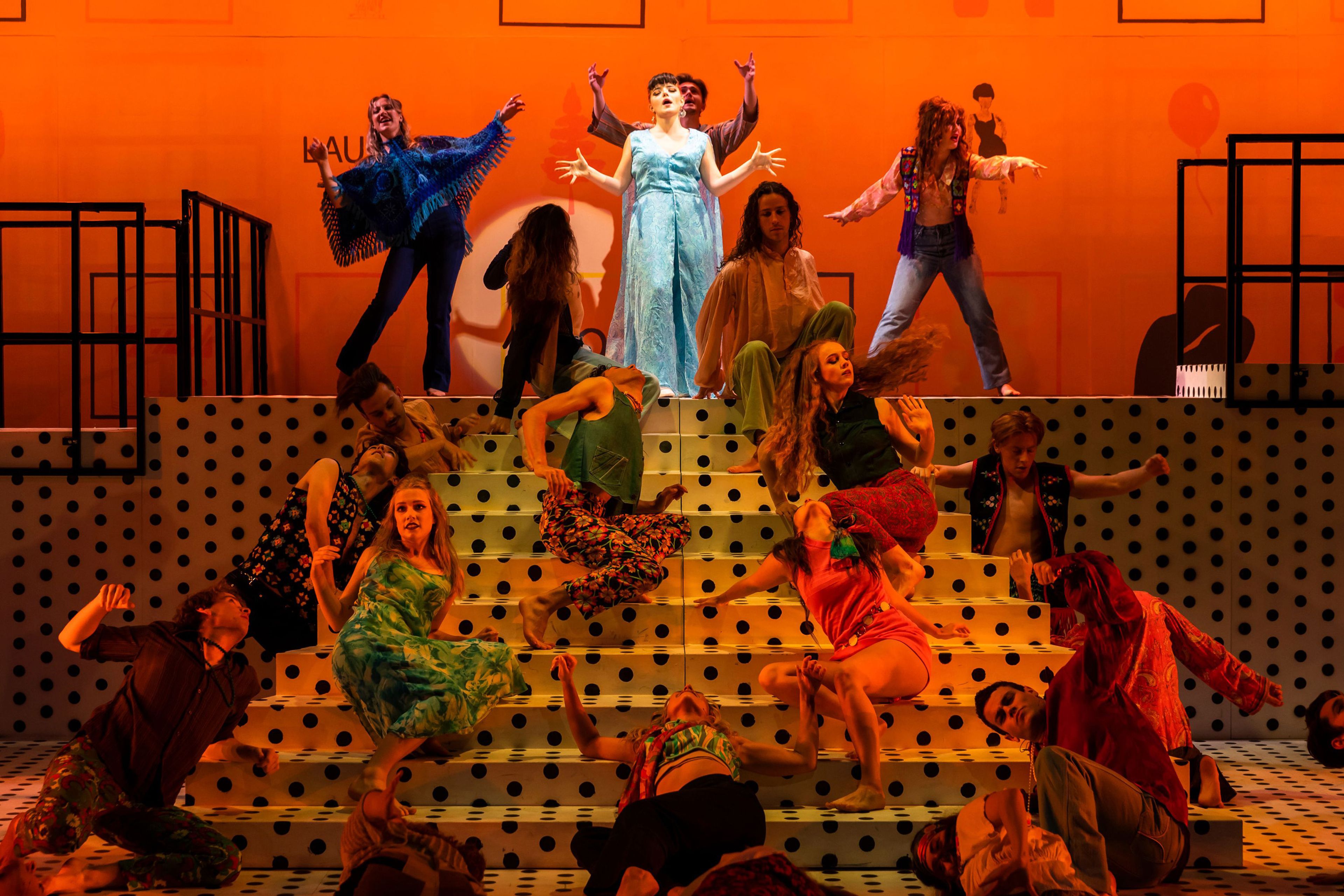 A vibrant theatrical performance scene featuring a central figure in a light blue dress standing on polka-dotted stairs, surrounded by energetic dancers in colourful, retro-inspired outfits