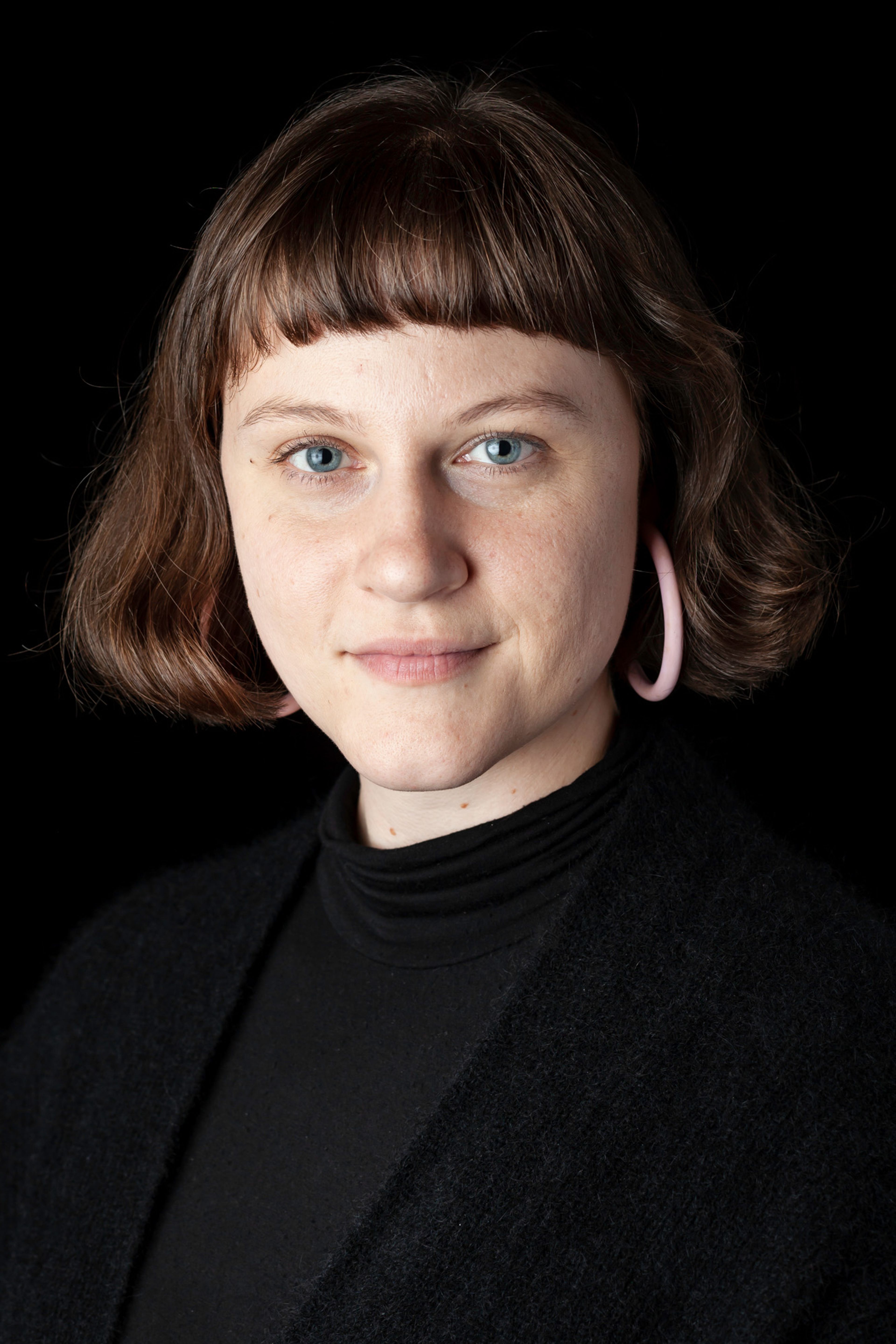 A portrait of a person with short brown hair and blue eyes, wearing a black turtleneck and large circular pink earrings