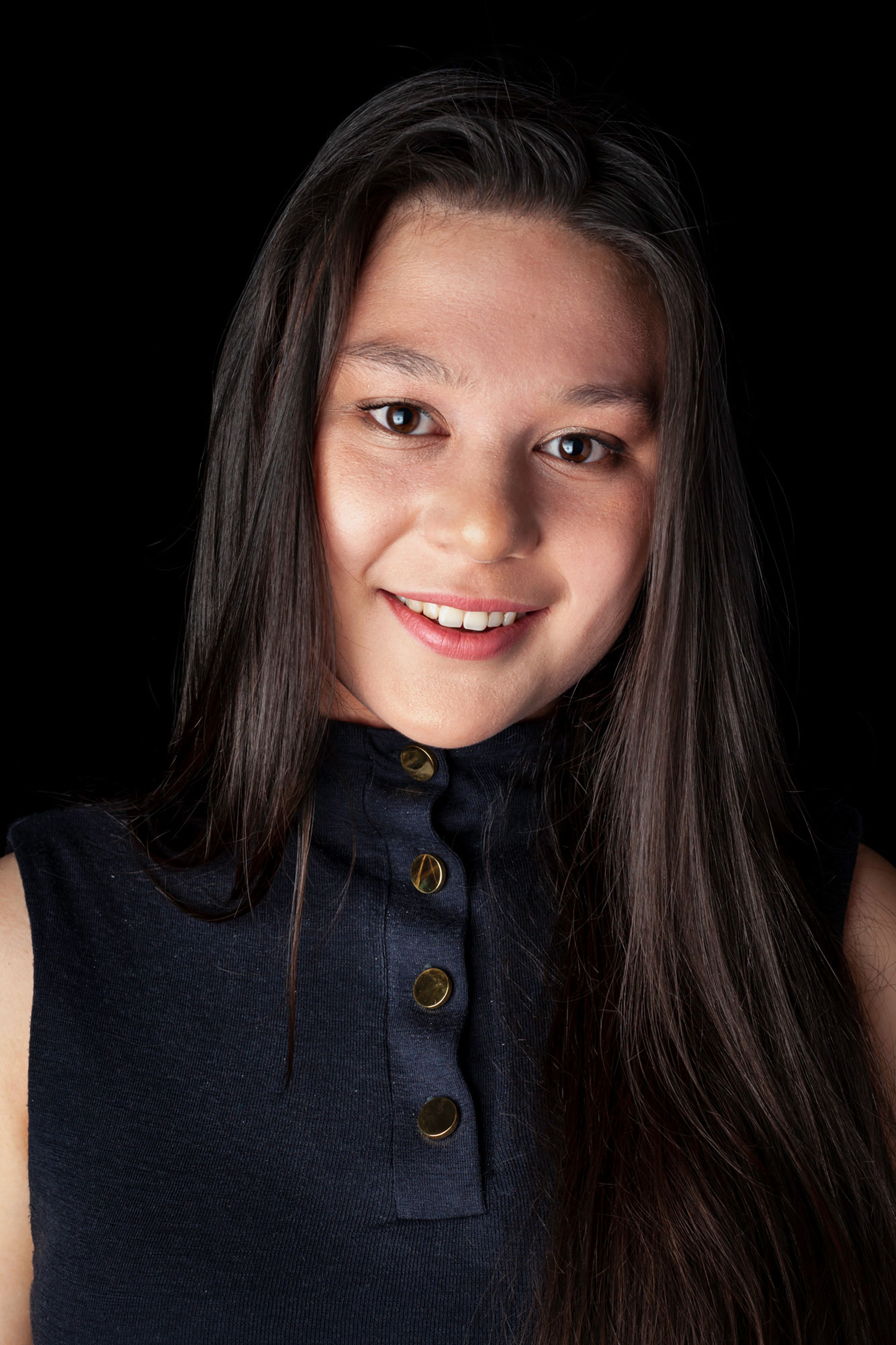 A portrait of a young woman with long, straight dark hair and a warm smile, wearing a sleeveless navy top with gold buttons