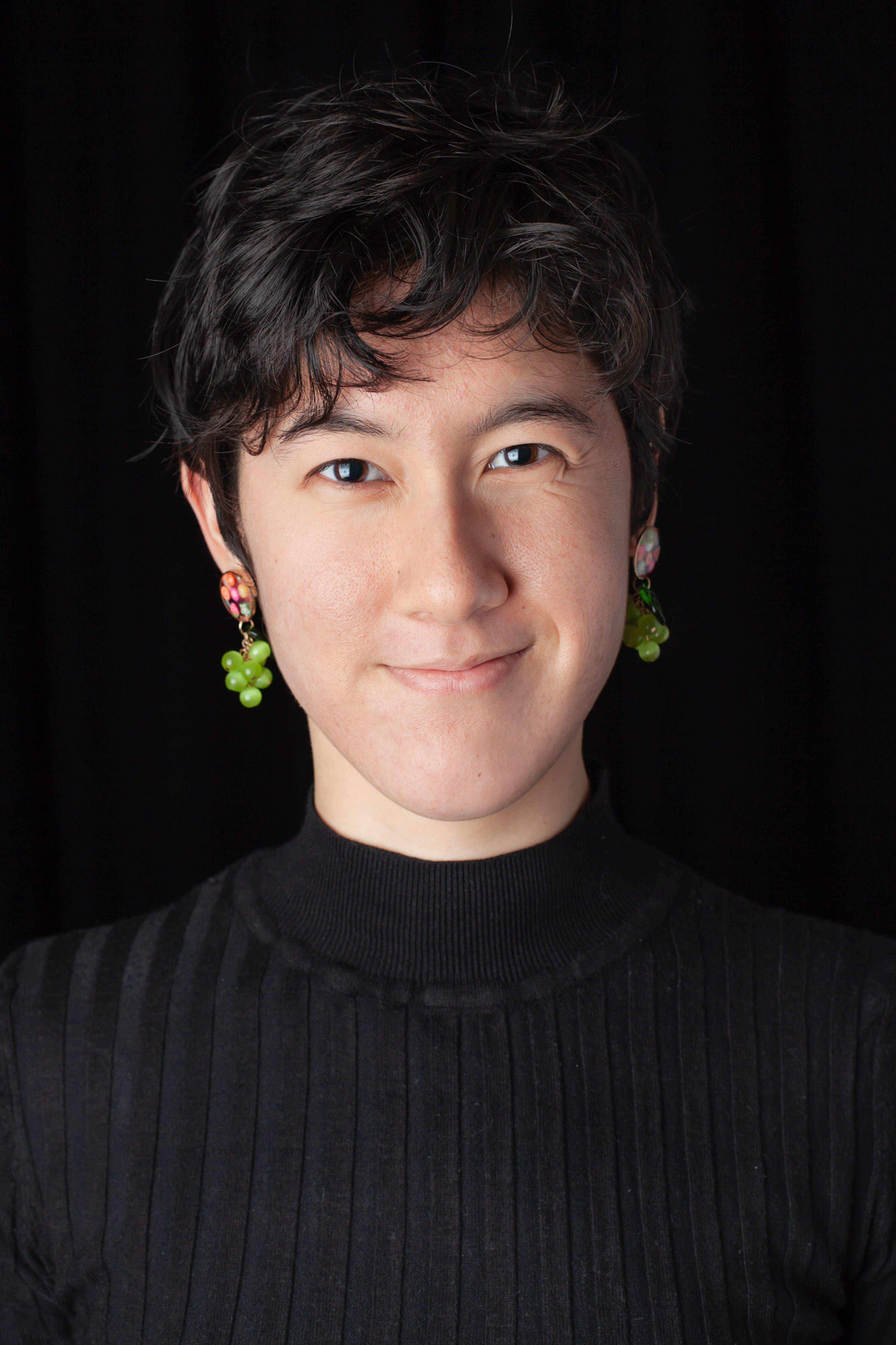 A portrait of a person wearing a ribbed black turtleneck and statement earrings resembling small green grapes, they have short, tousled hair