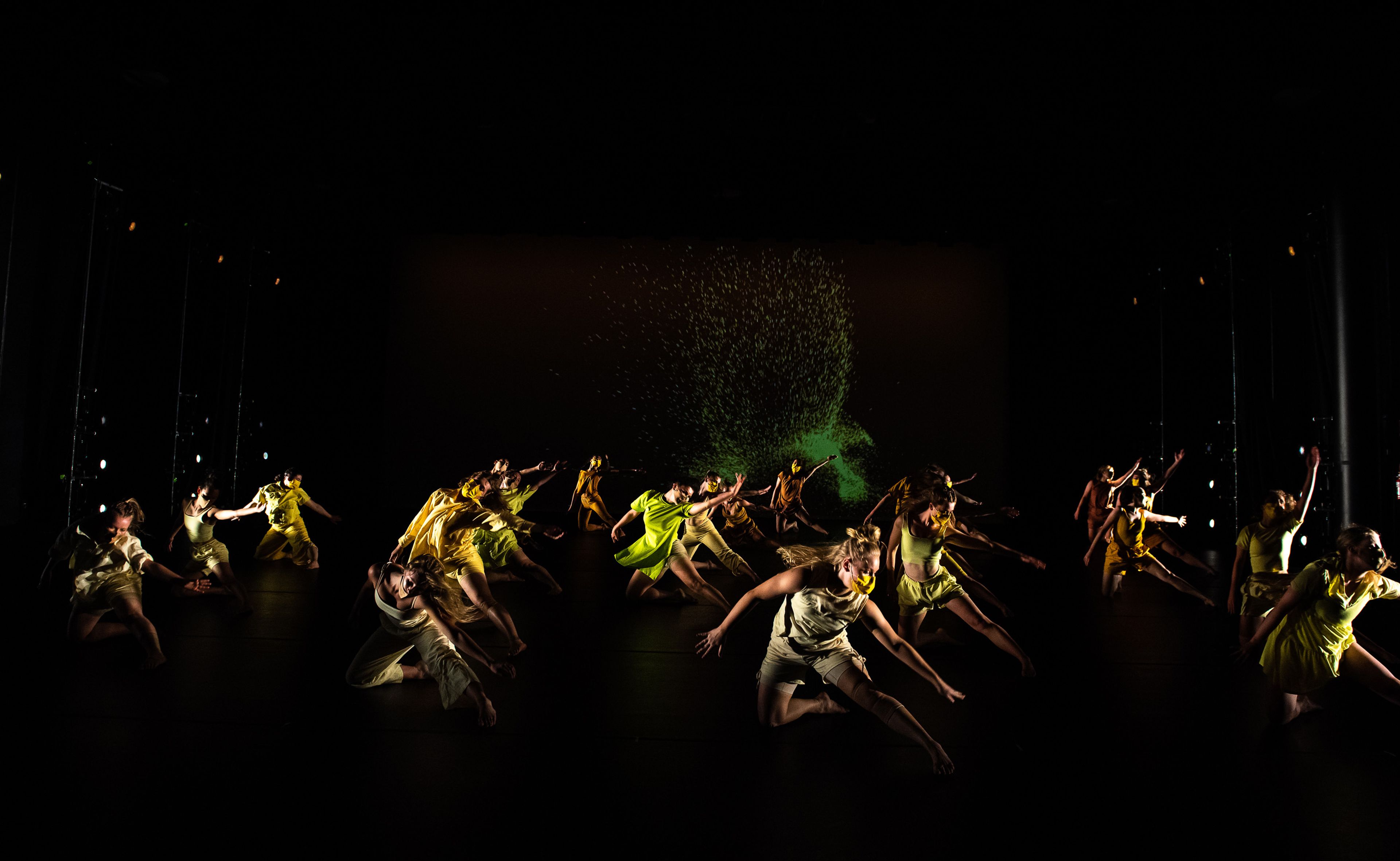 Dancers in yellow costumes captured mid-motion on a dimly lit stage with green projection in background