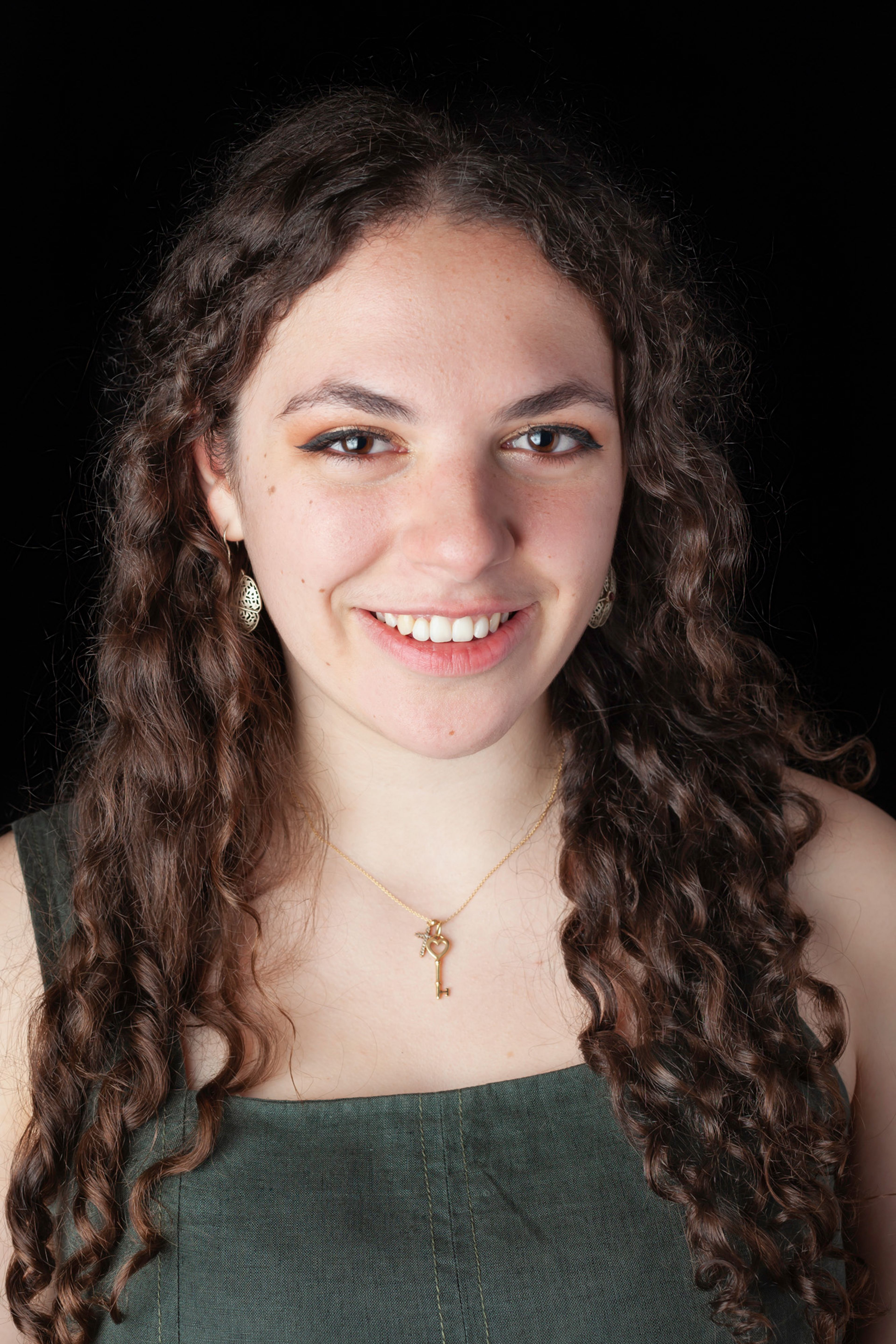 Portrait of a person with long, dark curly hair wearing a green sleeveless top, gold earrings, and a gold key pendant necklace