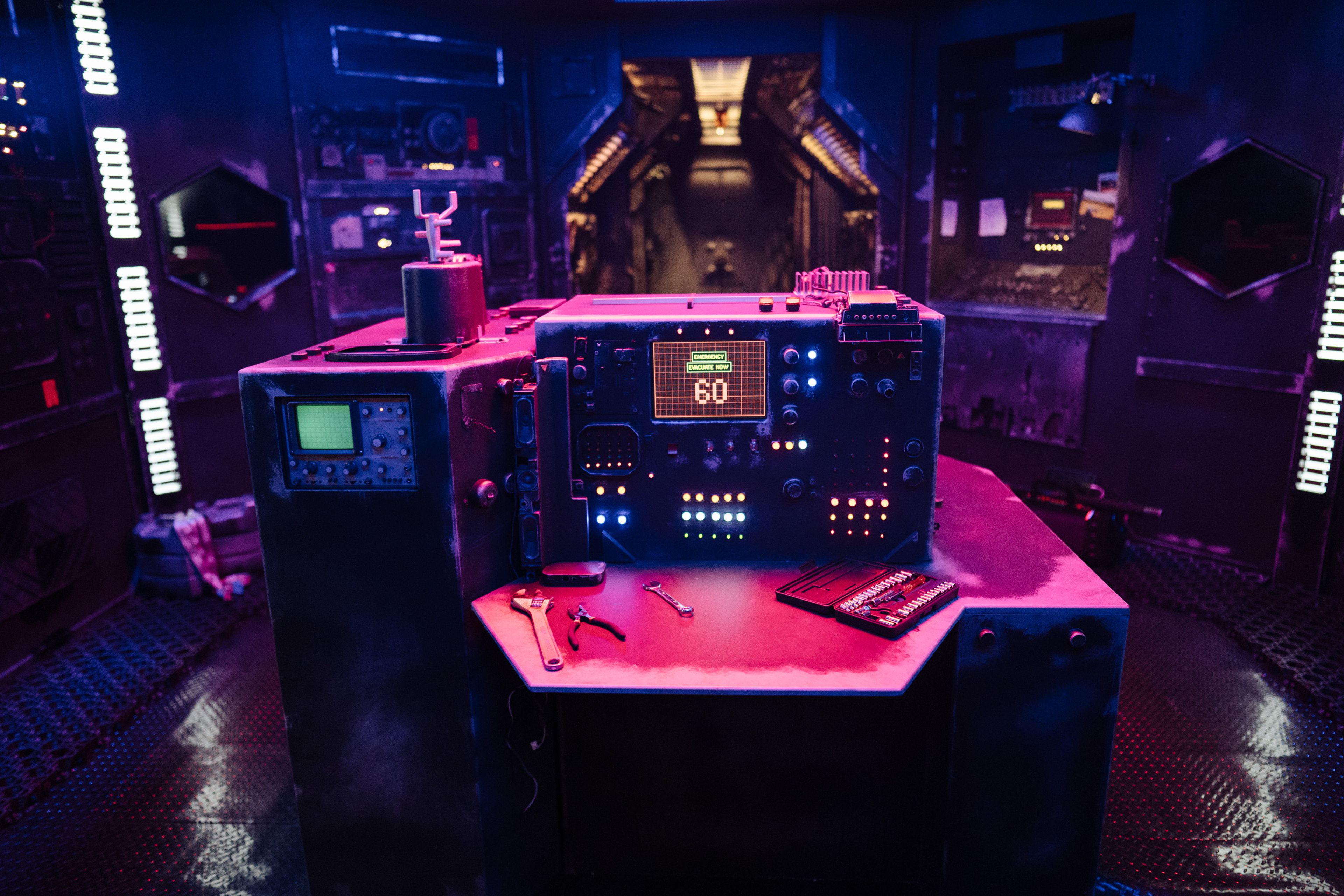 A sci-fi set featuring a control station with illuminated buttons, dials, and tools, bathed in dramatic blue and pink lighting, with a futuristic corridor in the background