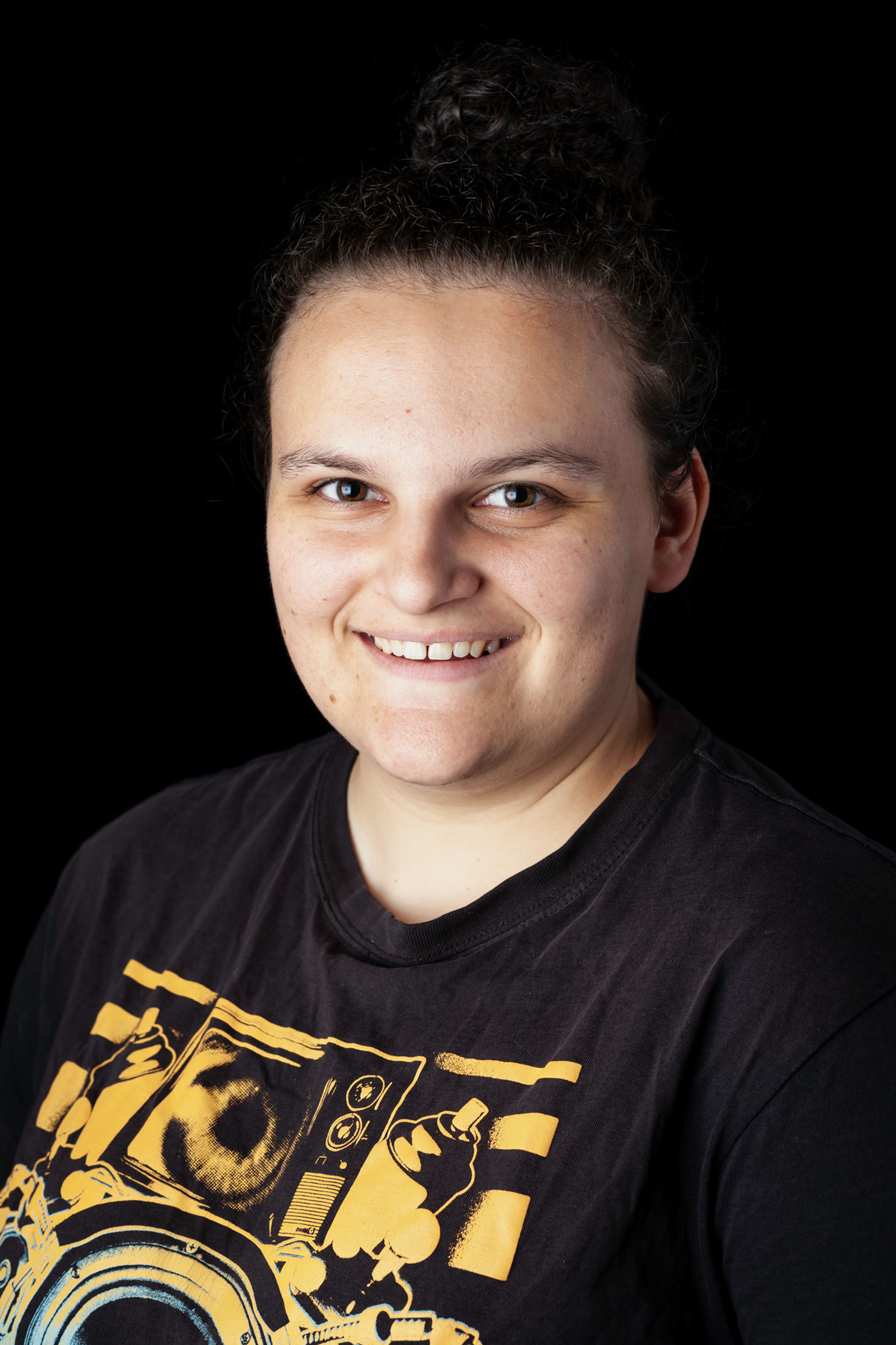 A portrait of a smiling individual with curly hair tied back, wearing a black graphic T-shirt featuring bold yellow imagery