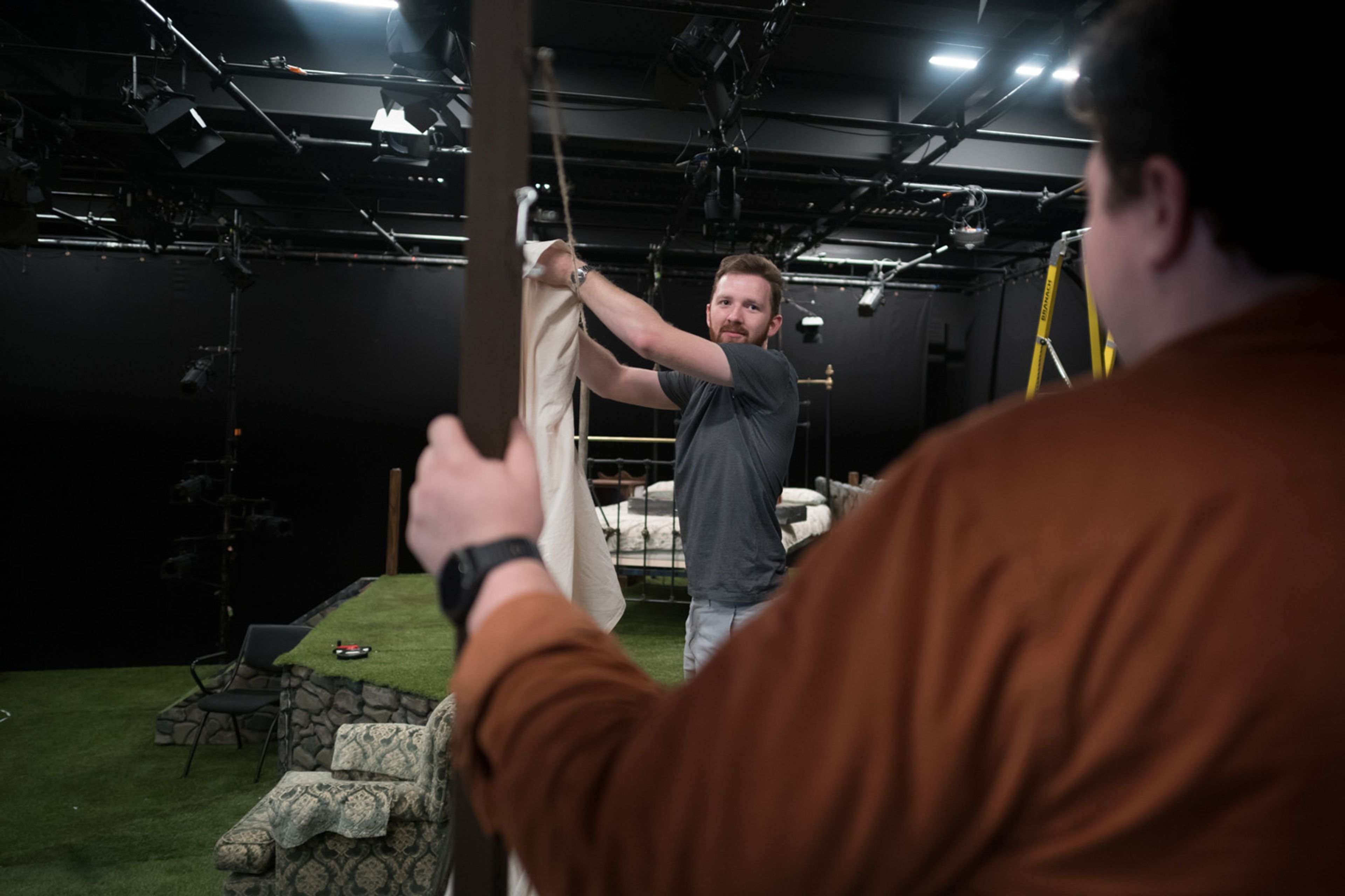 Two individuals working in a theatre. One, in the foreground wearing a brown shirt, holds a structure, while the other, in a grey t-shirt, adjusts a white fabric on a frame. The background features stage lighting, props, and a grassy platform