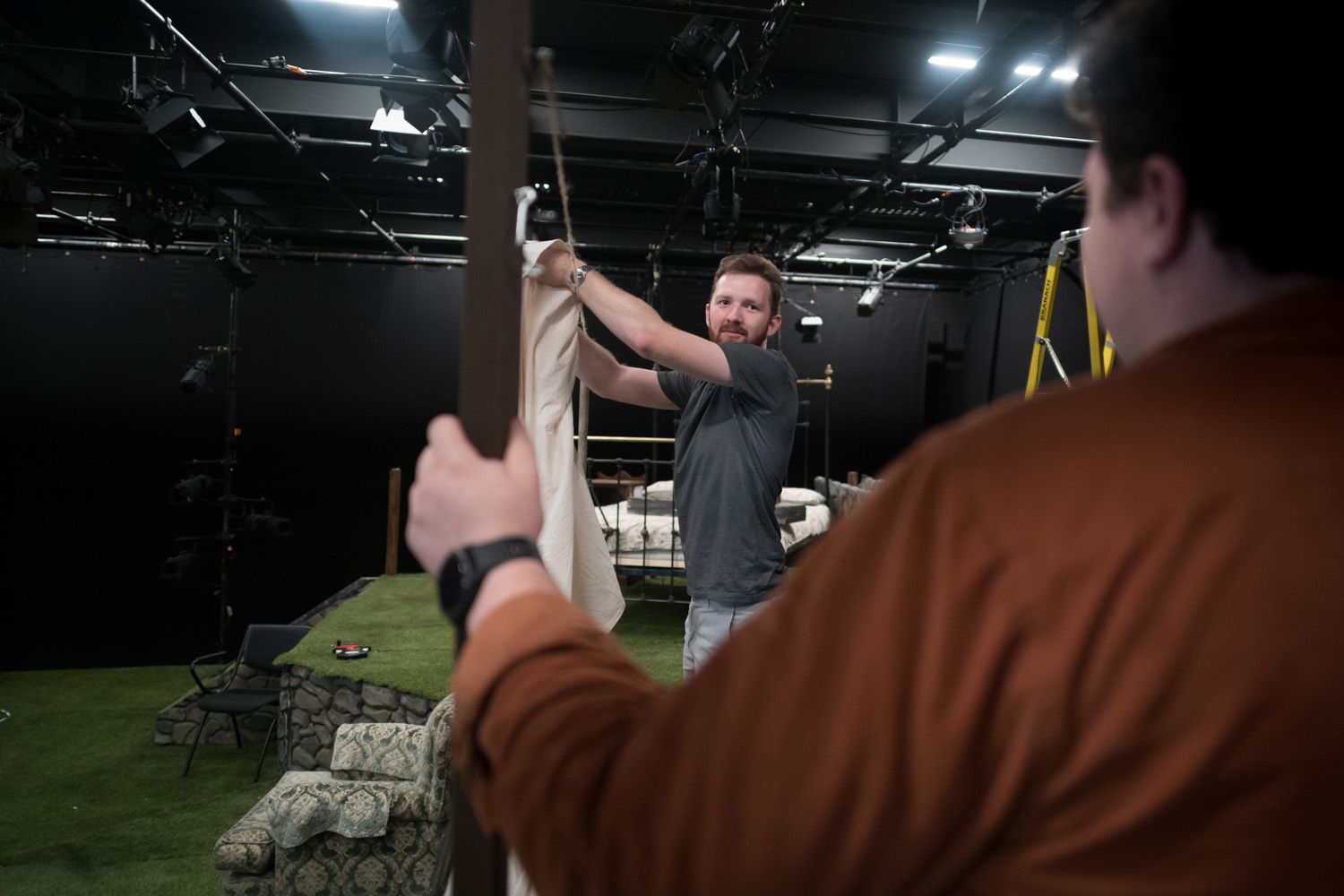 Two individuals working in a theatre. One, in the foreground wearing a brown shirt, holds a structure, while the other, in a grey t-shirt, adjusts a white fabric on a frame. The background features stage lighting, props, and a grassy platform