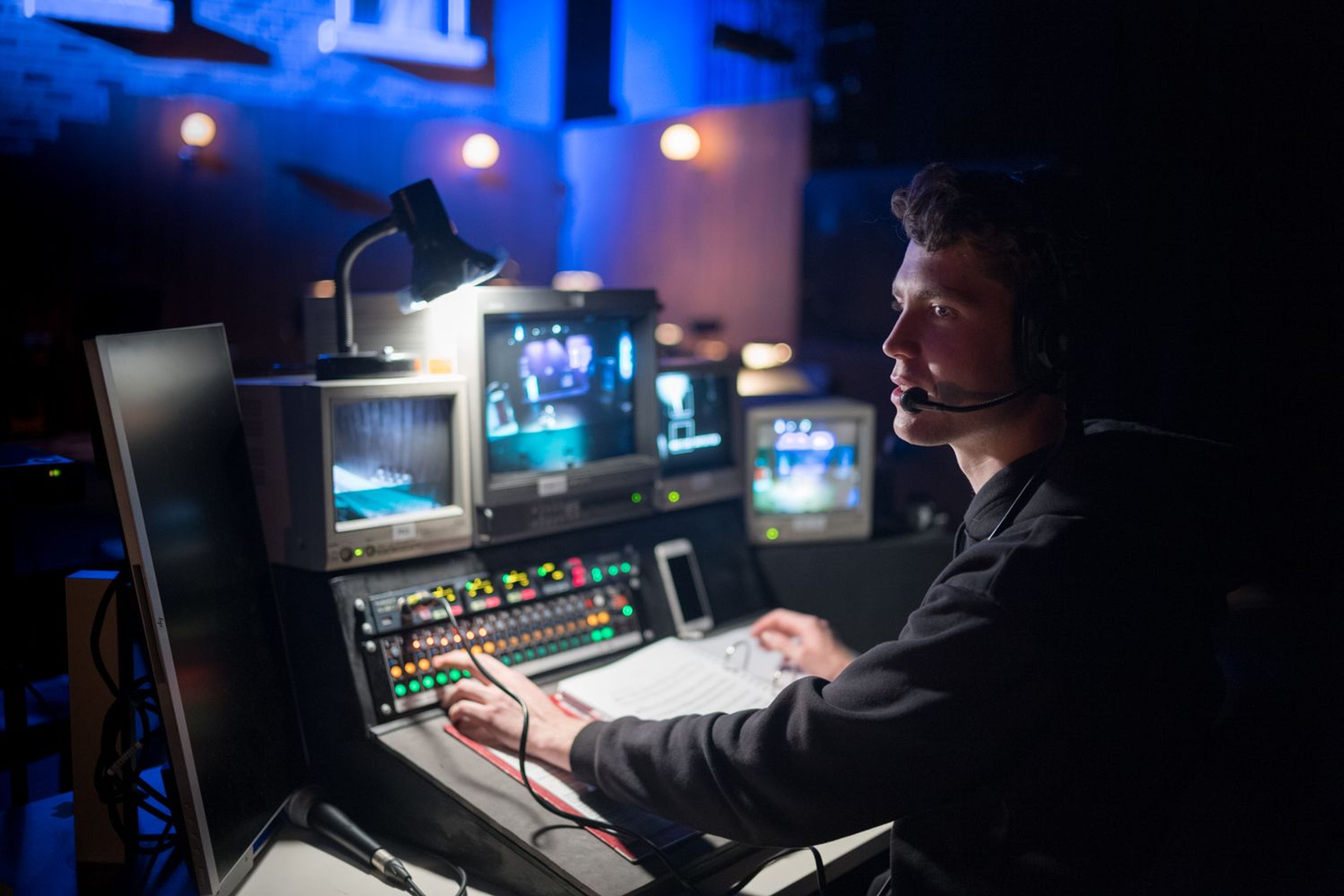 A person wearing a headset operates a control desk in a dimly lit theatre with multiple monitors displaying video from stage