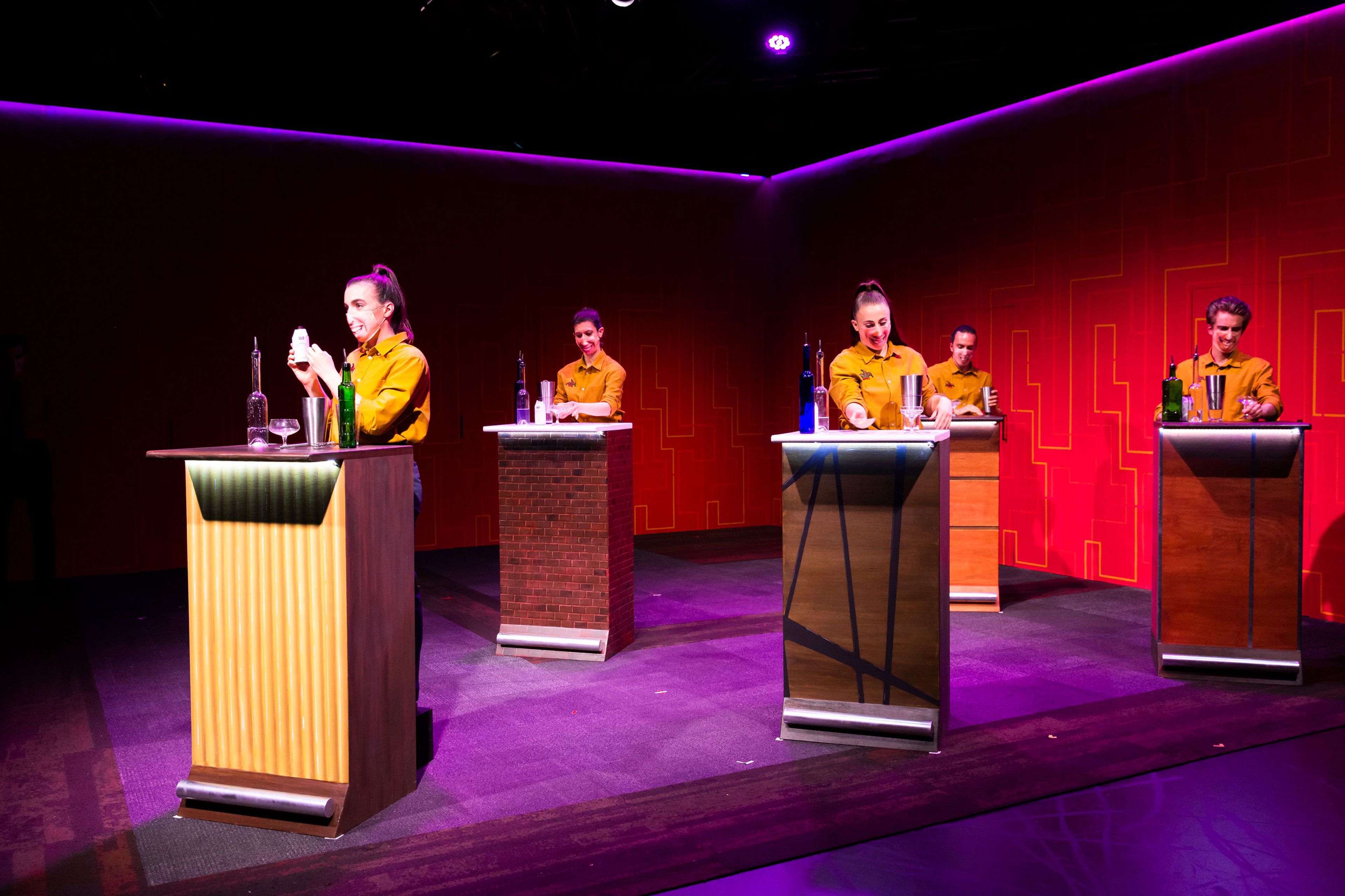 The image shows five performers in mustard uniforms behind podiums, engaged in bartending, against a red geometric backdrop with pink and purple lighting