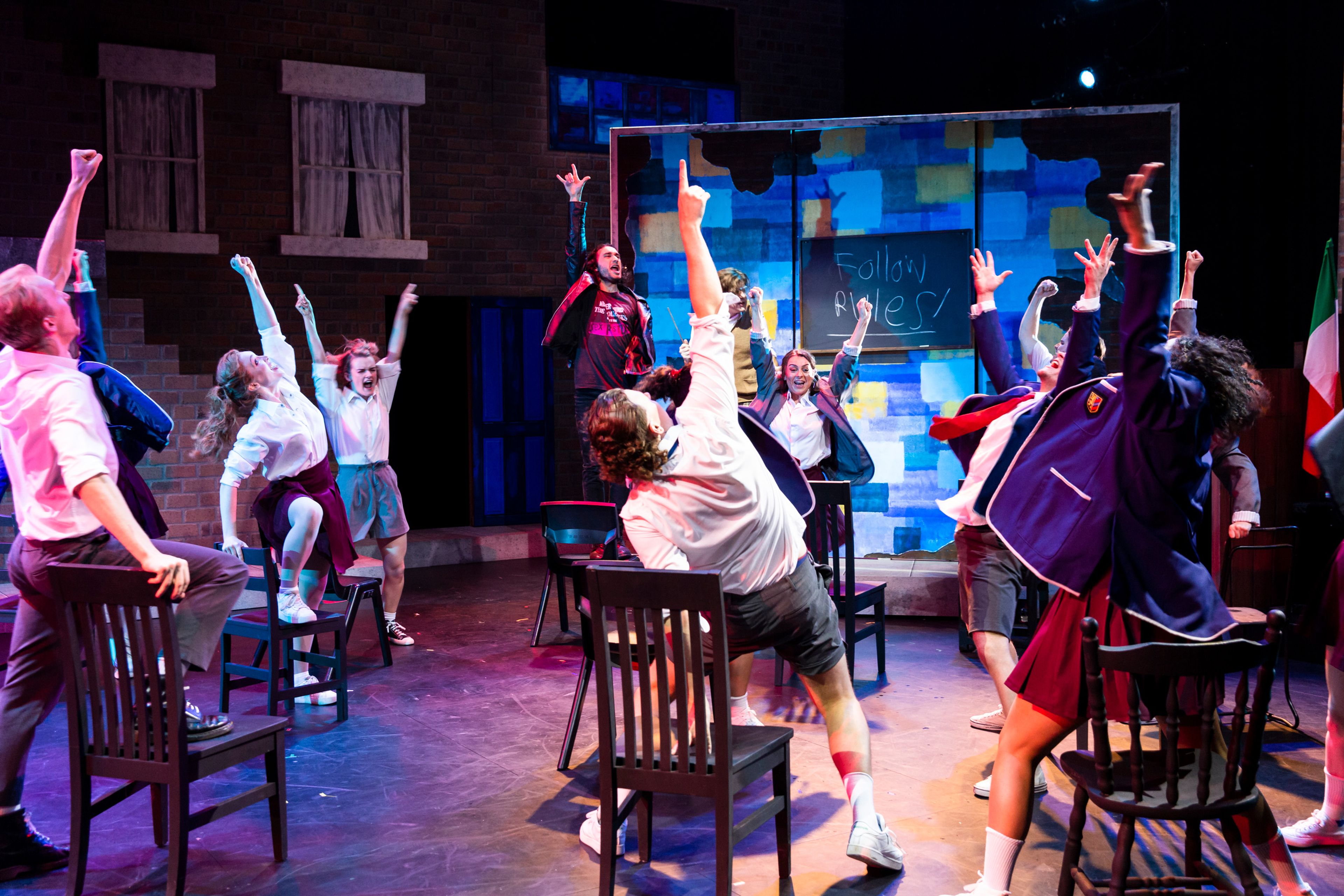 Performers posing energetically in school costumes on chairs against a backdrop of brick walls and a colorful chalkboard reading "Follow Rules!"