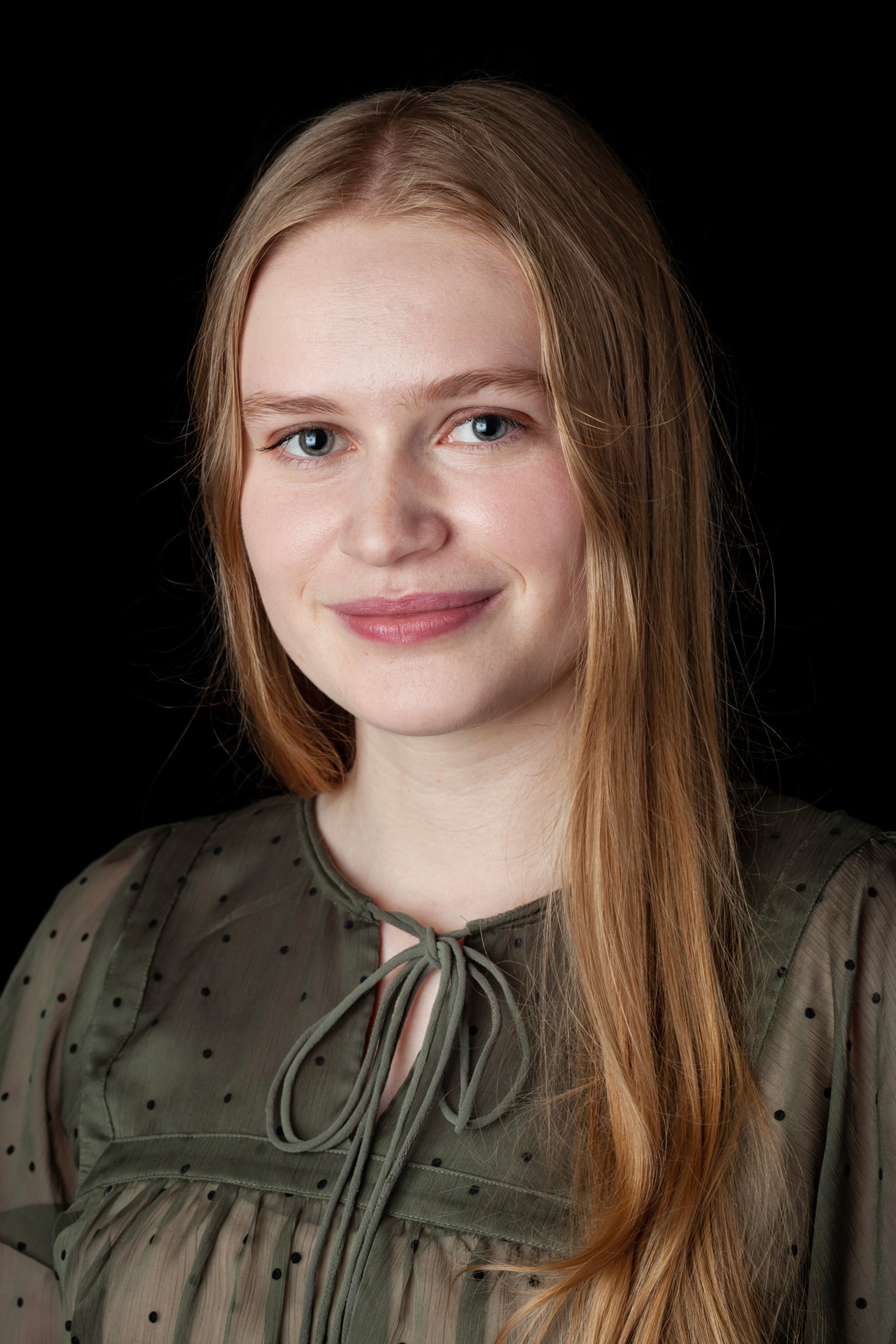 A portrait of a young woman with long blonde hair, wearing a sheer green blouse adorned with small black polka dots and tied at the neck