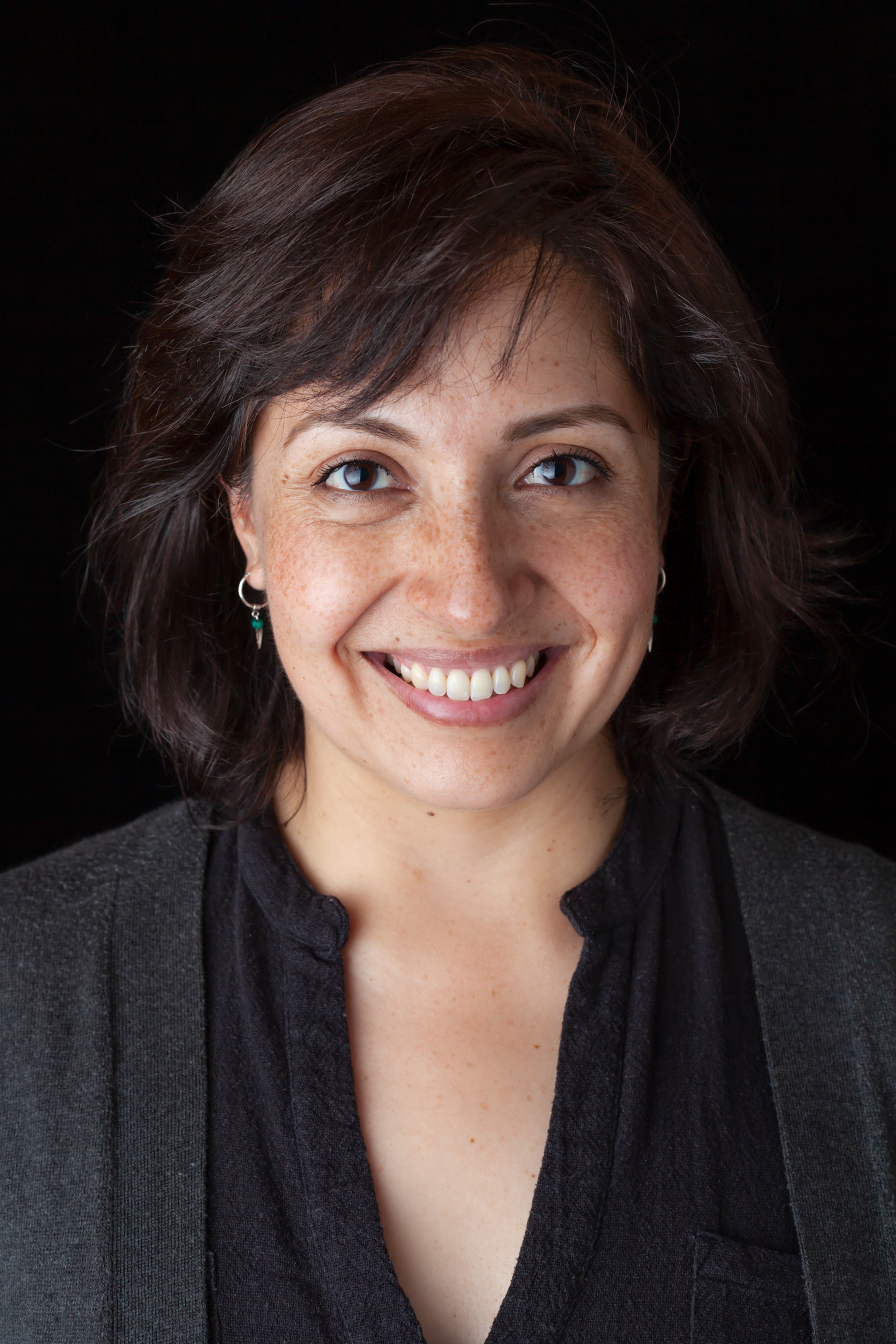 A portrait of a person with short, dark hair, smiling warmly, wearing a dark top and cardigan, with subtle freckles and small earrings