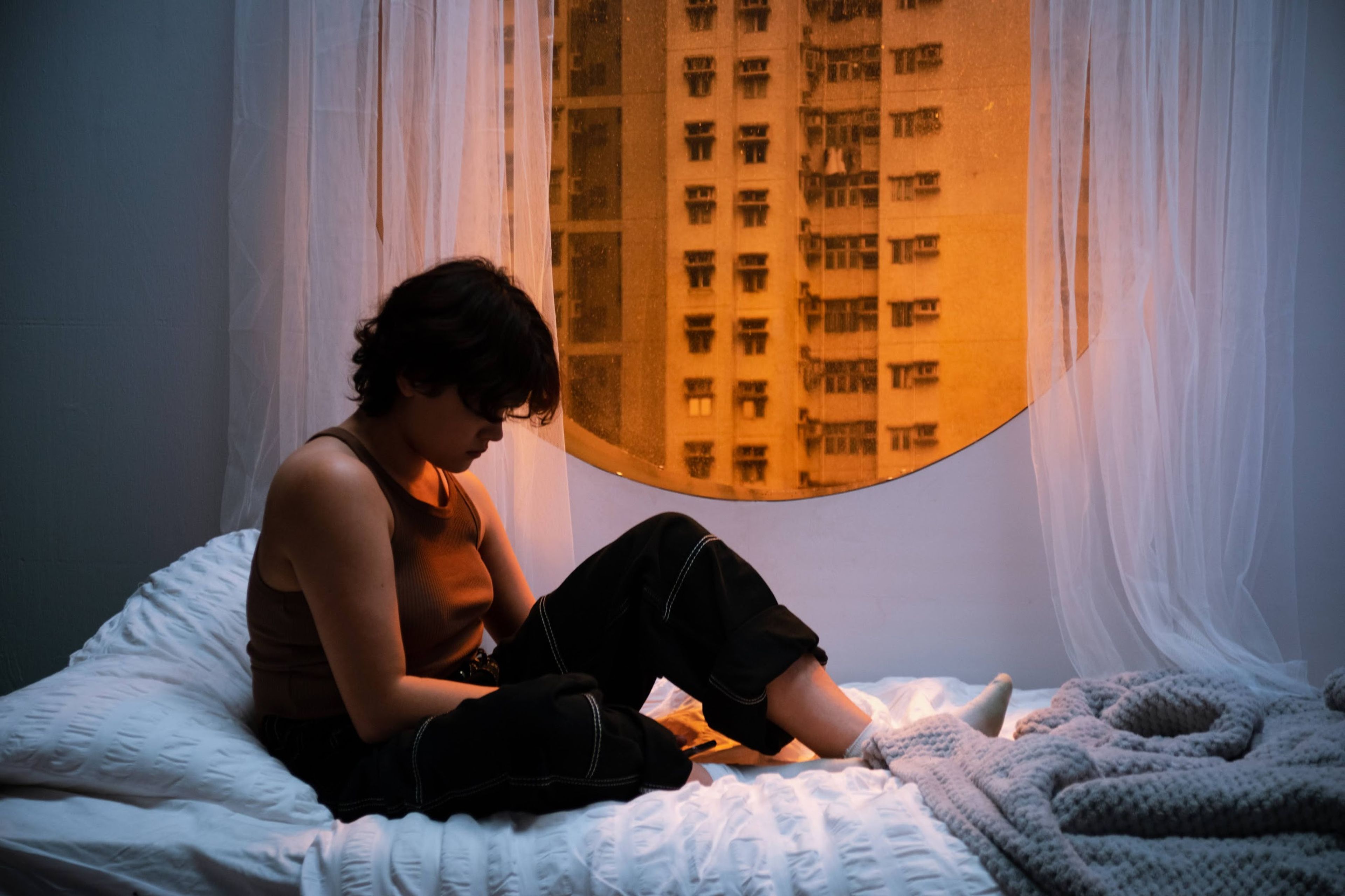 a person sitting on a bed near a circular window. Warm orange light from a cityscape filters through the window, contrasting with the cool tones of the interior