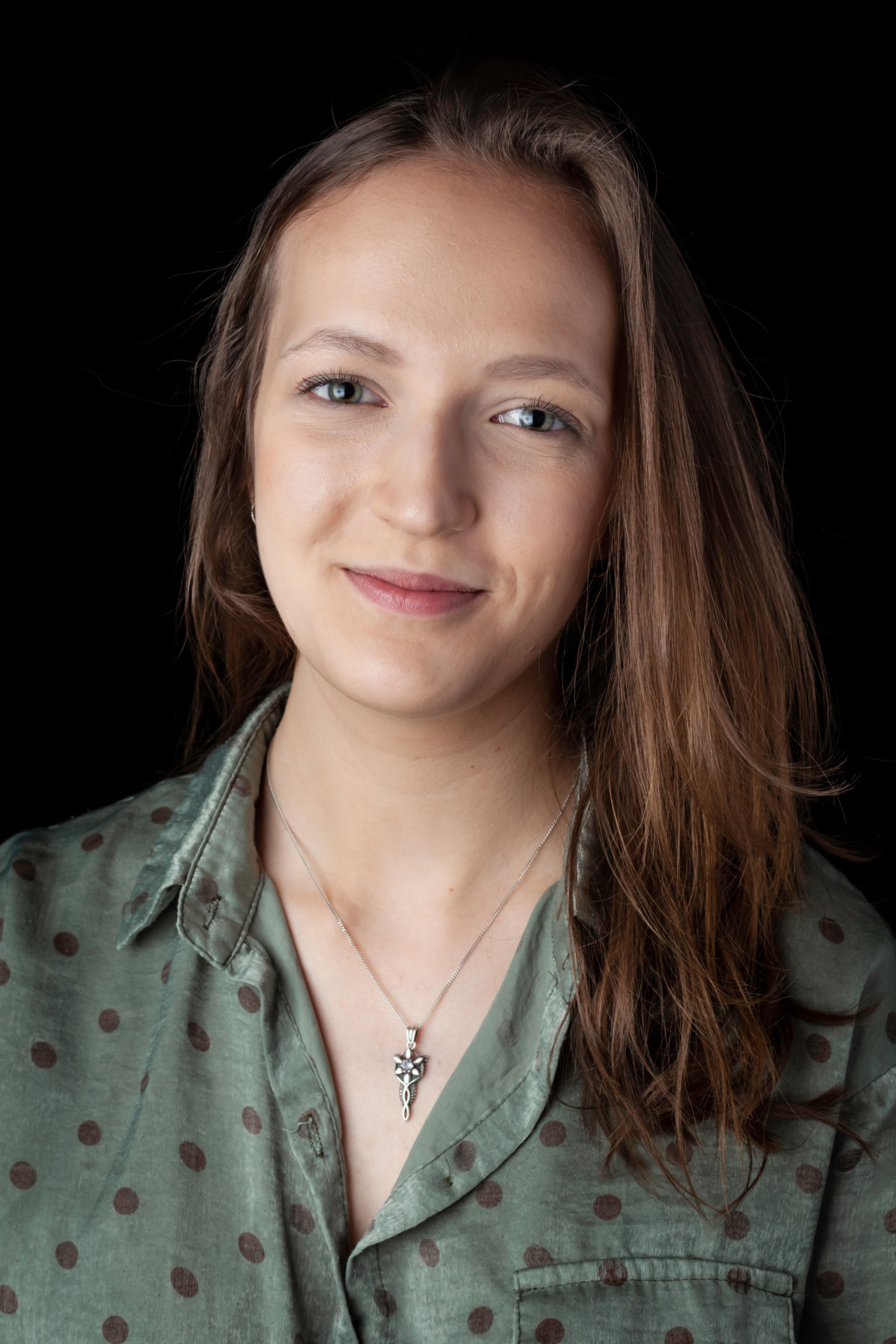 This is a headshot of a woman with long brown hair, wearing a green polka-dot shirt and a delicate silver pendant necklace