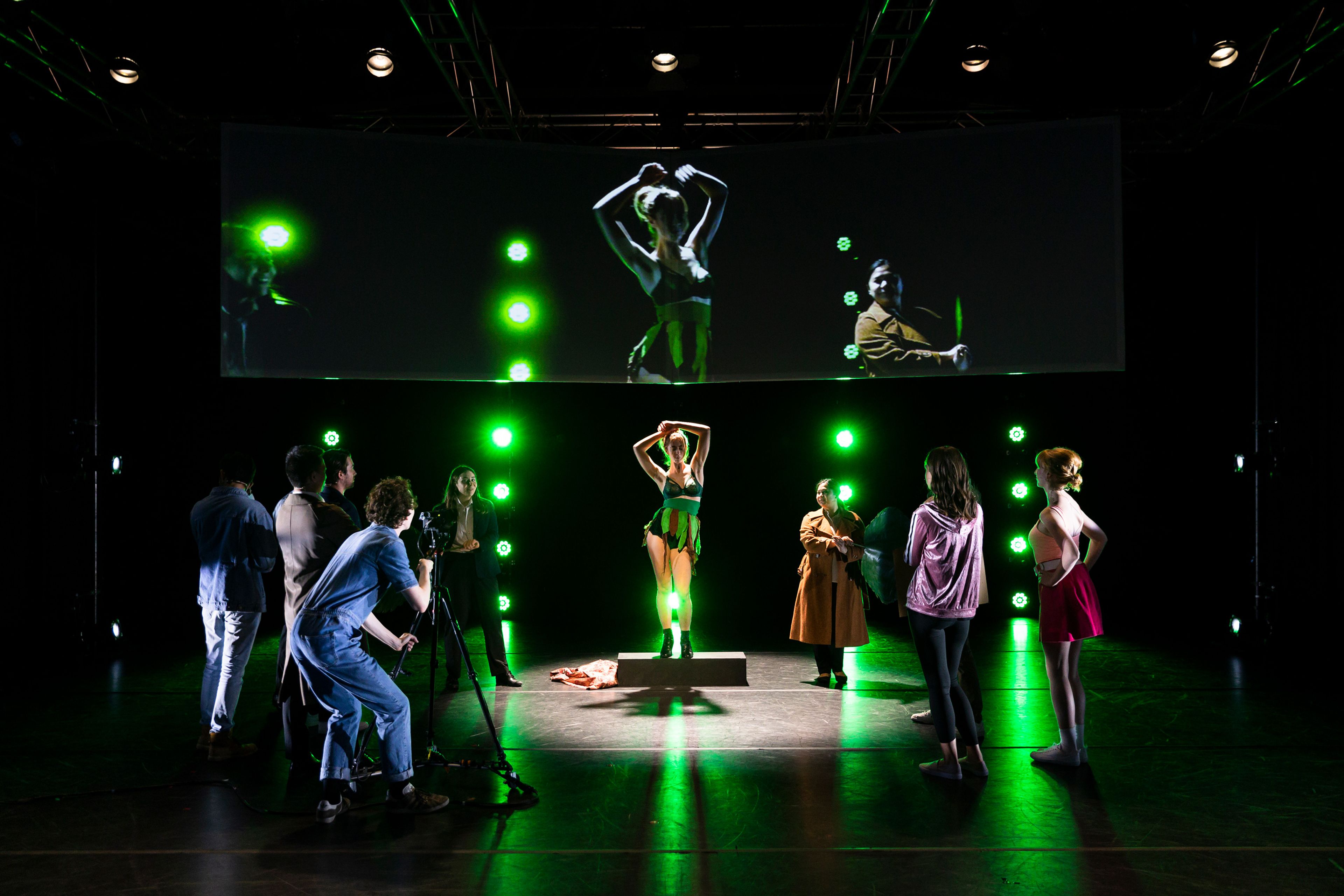 A theatrical scene featuring a performer in a green costume on a raised platform, illuminated by green stage lights