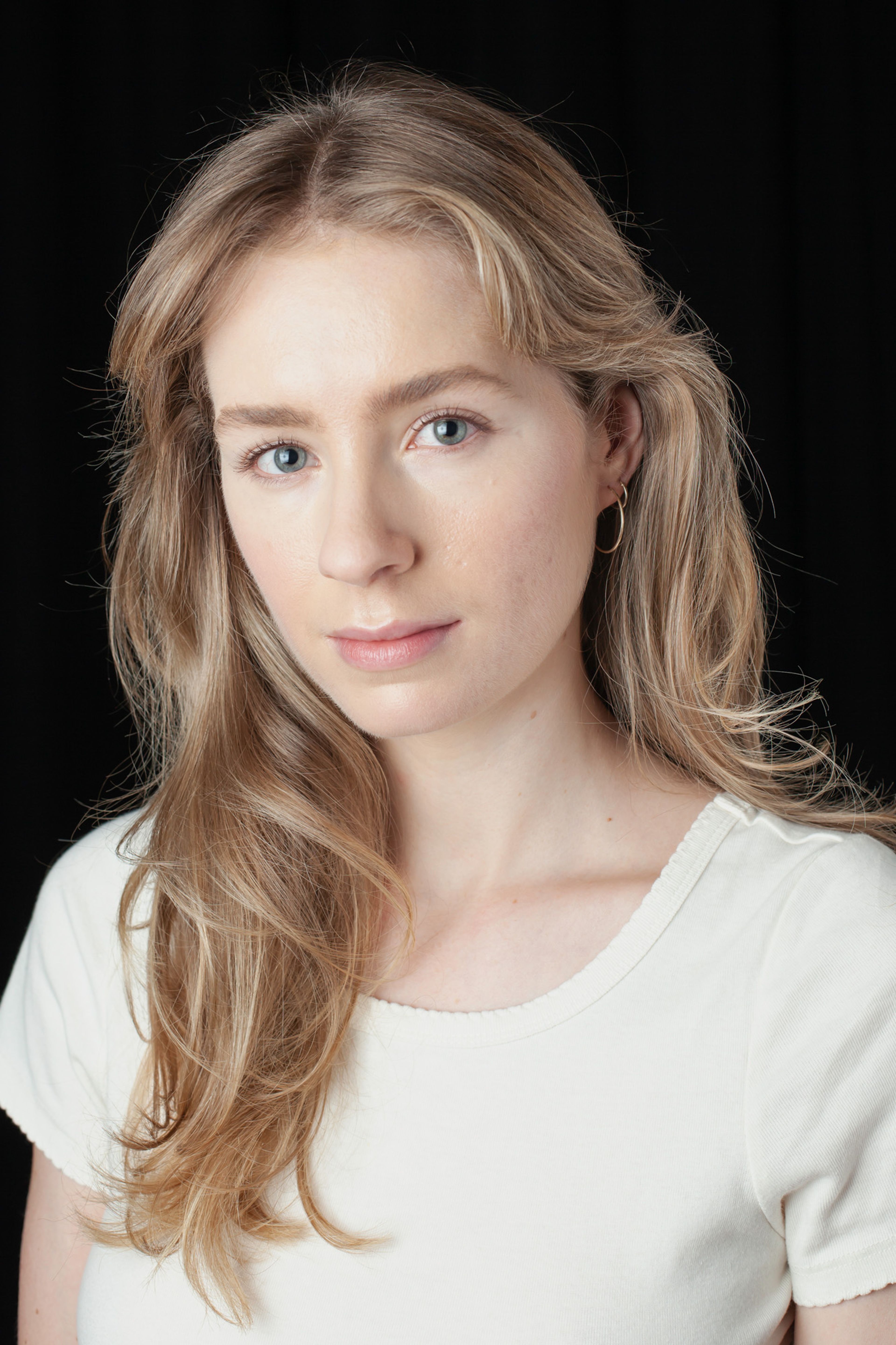 Portrait of a young woman with light blonde hair, styled in loose waves, wearing a simple white top and a delicate gold hoop earring