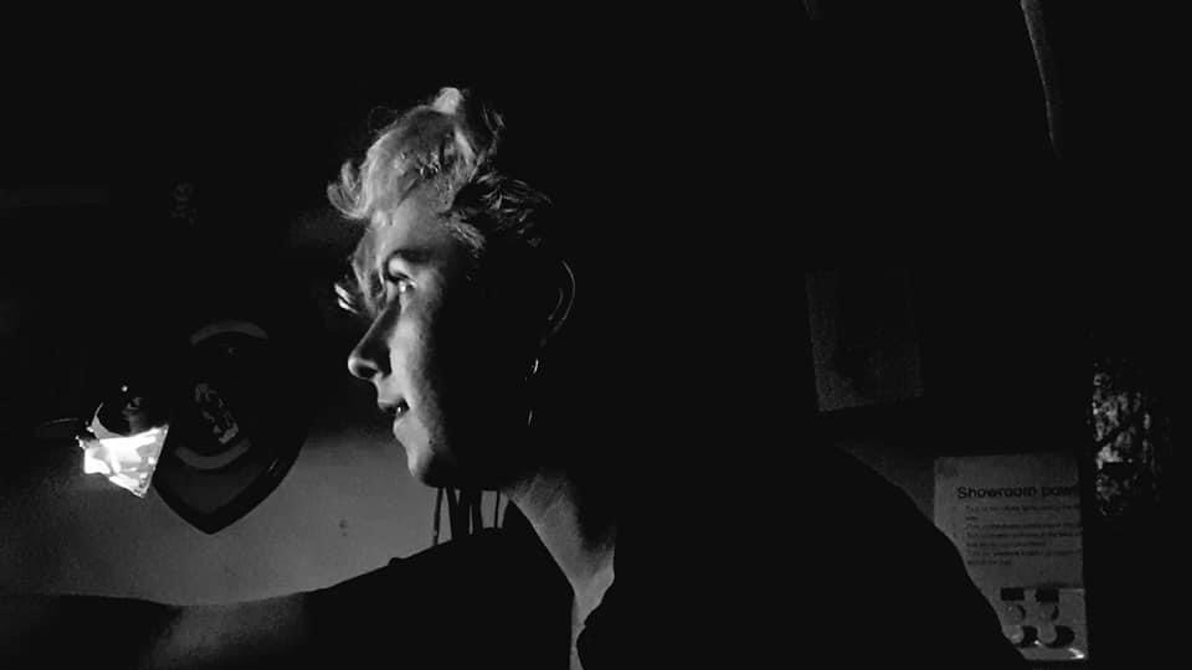 Black-and-white profile shot of a person with short, light-colored hair, illuminated by a small light source in a dark theatre