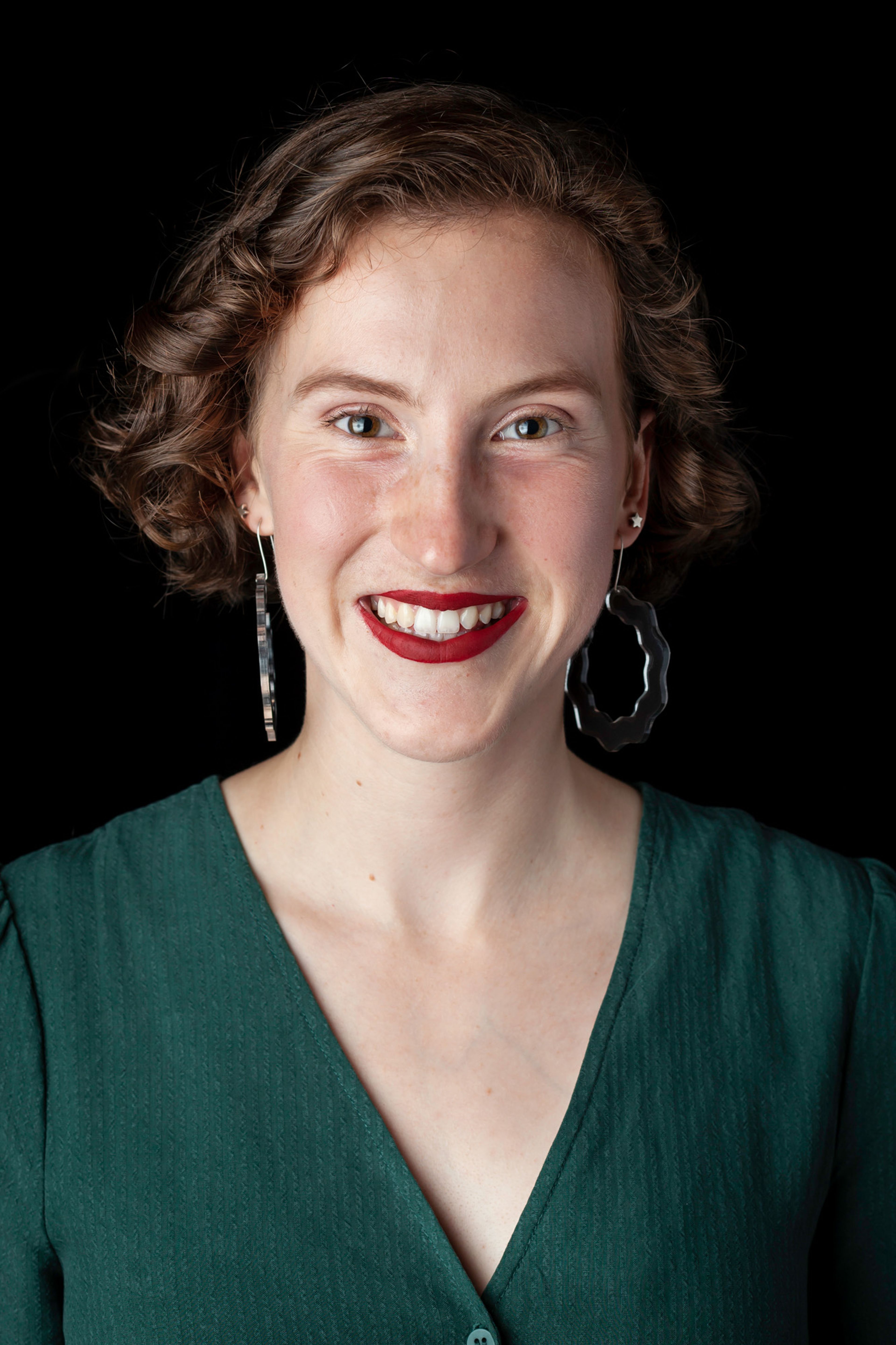 An individual with short, curly hair, wearing a green top and bold red lipstick. They have unique, wavy-shaped dangling earrings.