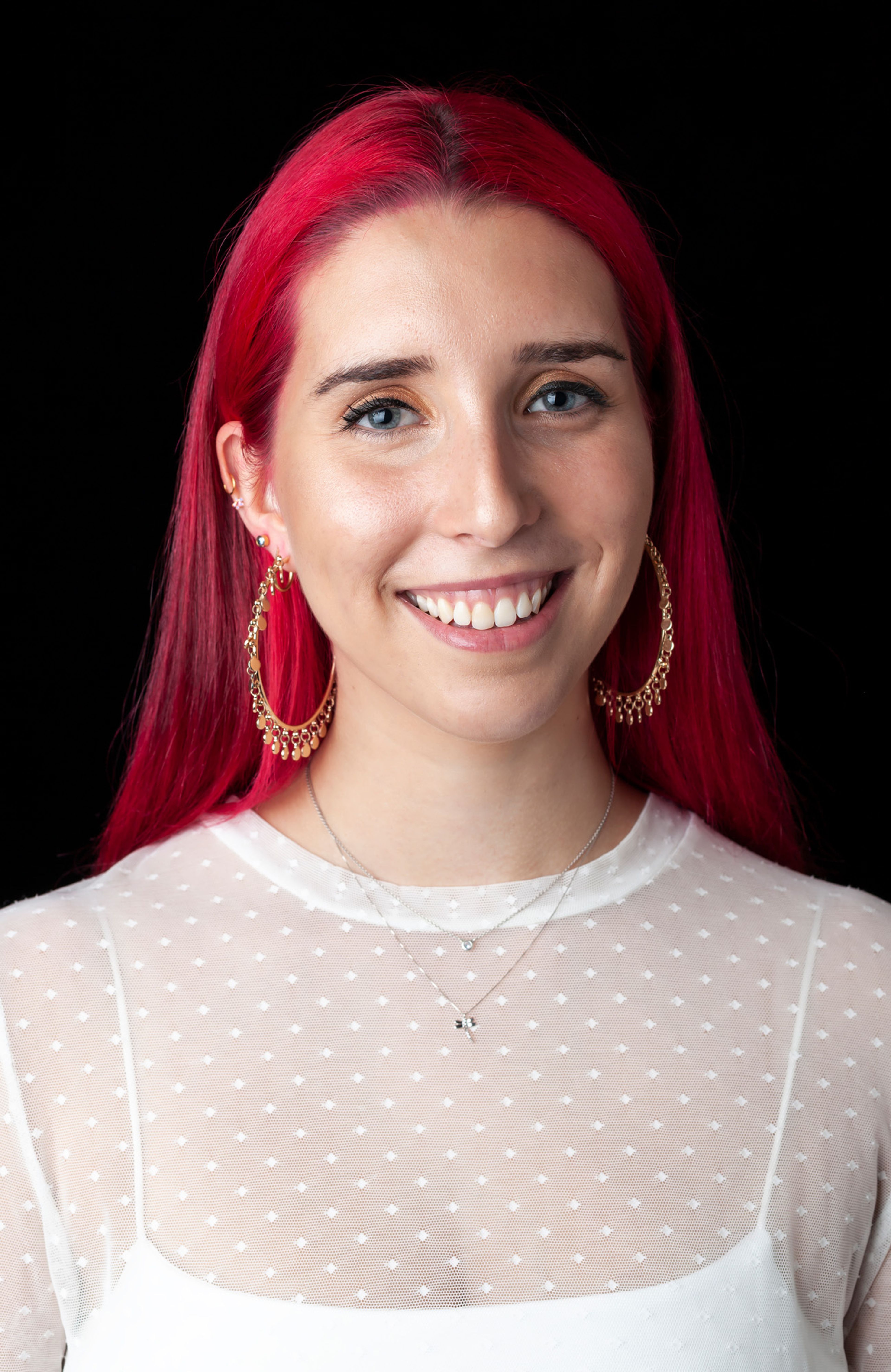 A smiling individual with vibrant red hair wears gold hoop earrings, a delicate necklace, and a sheer white top with a dotted pattern