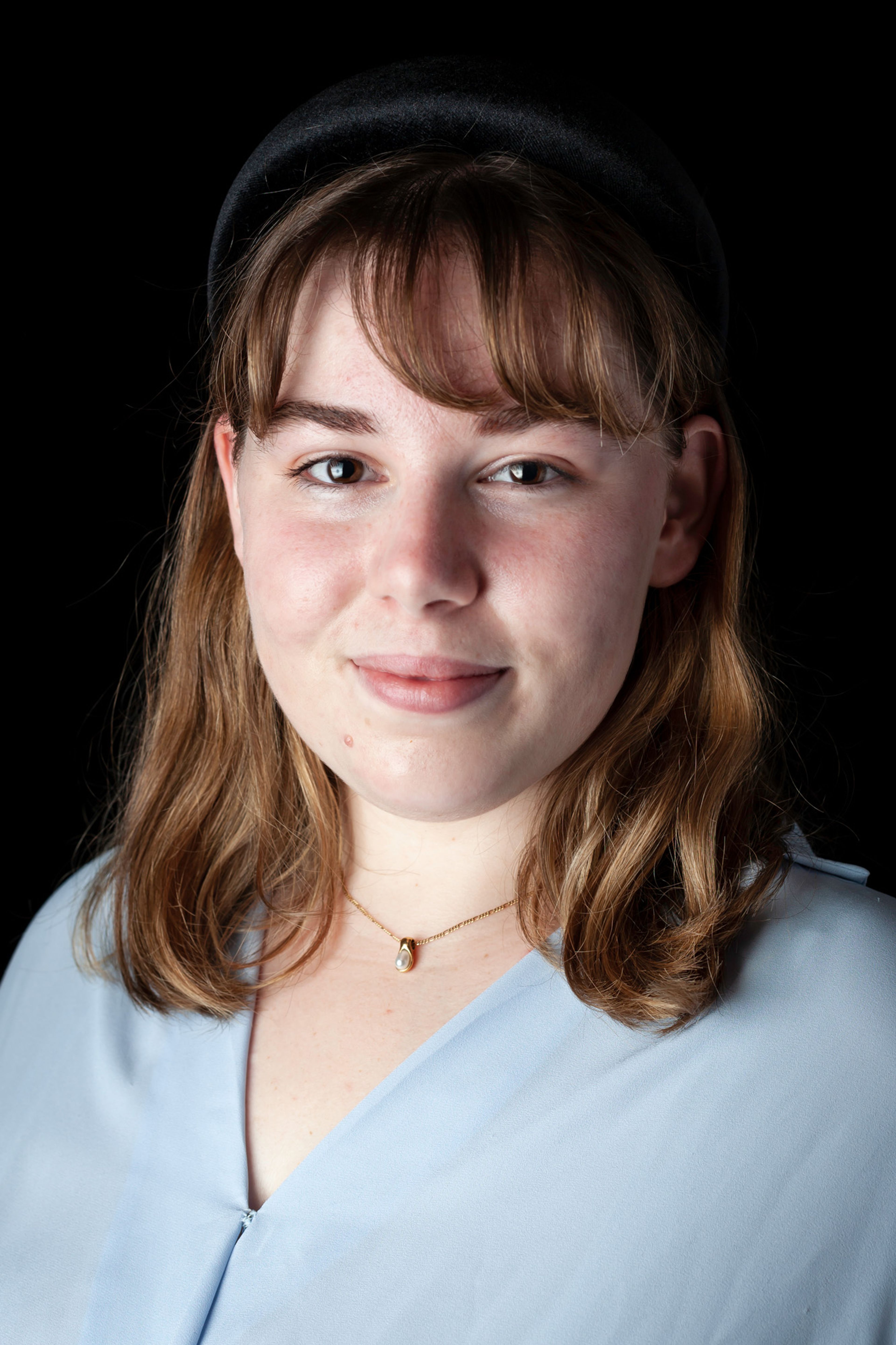 A portrait of a young woman with shoulder-length light brown hair, wearing a light blue blouse and a delicate gold necklace with a pearl pendant