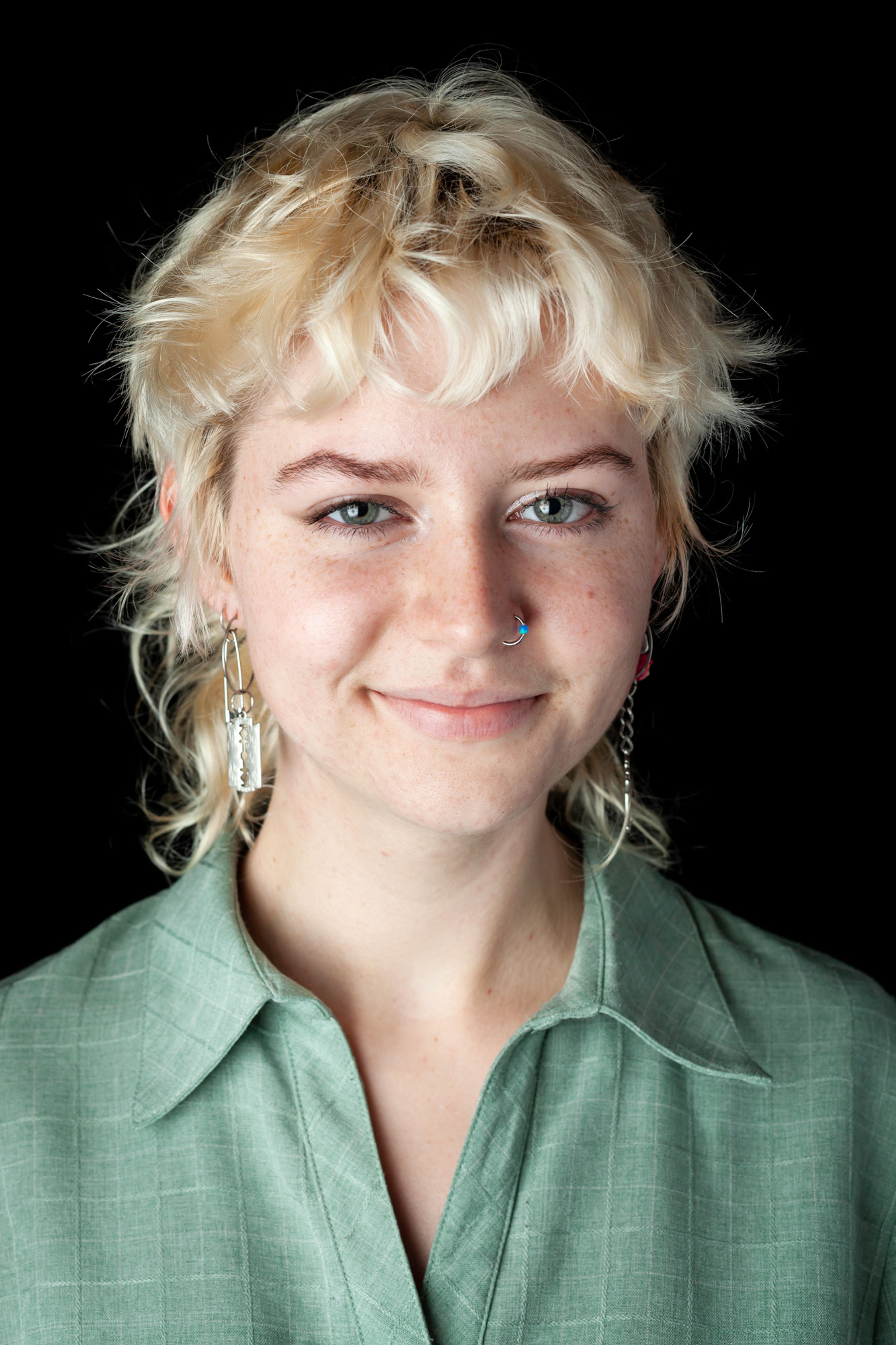 A portrait of a person with short, light blonde hair wearing a green collared shirt, silver dangling earrings, and a nose ring