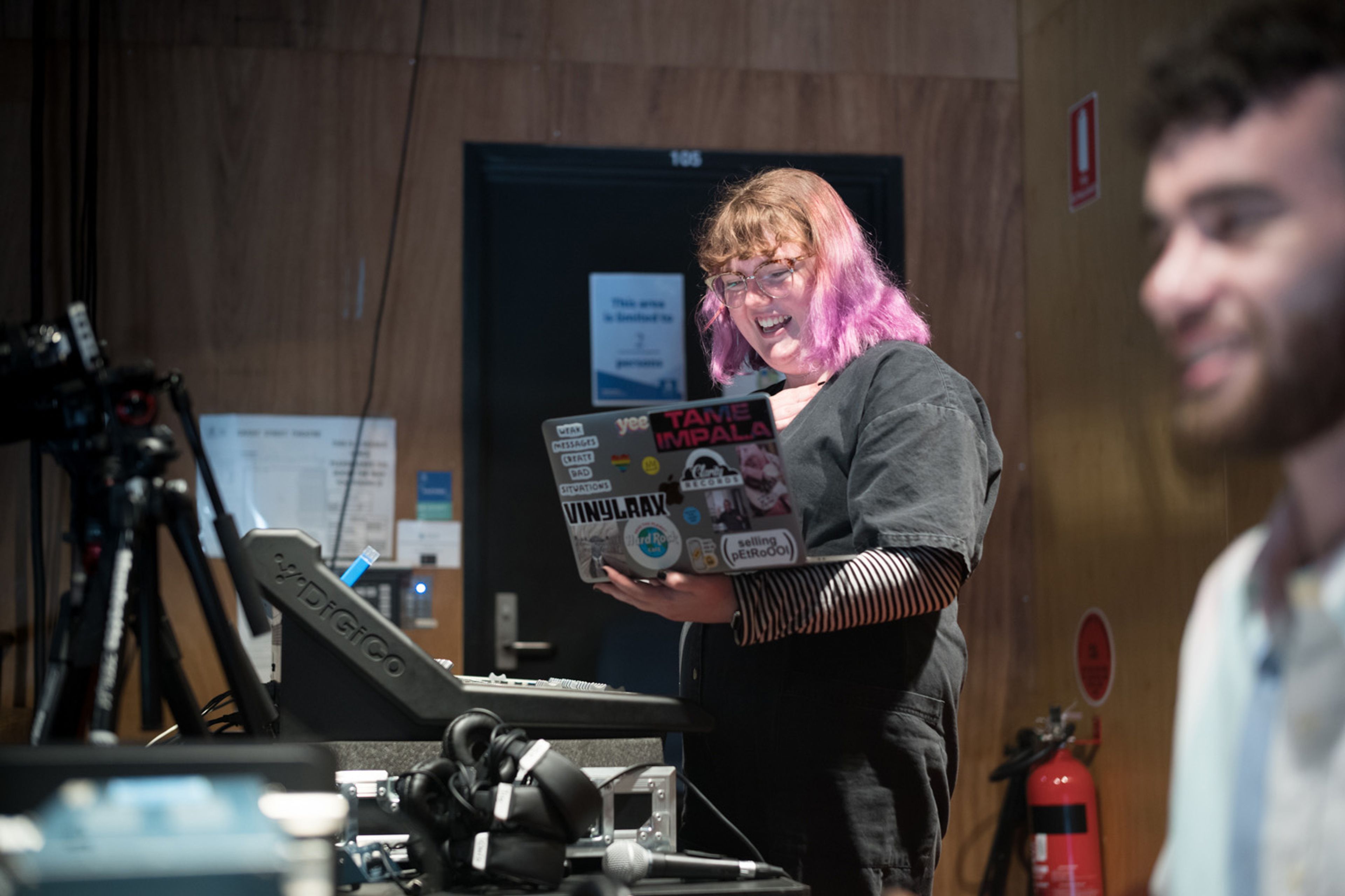 A person with purple hair, wearing glasses and a black outfit, smiles while working on a laptop covered in colorful stickers. They are surrounded by sound equipment in a control room