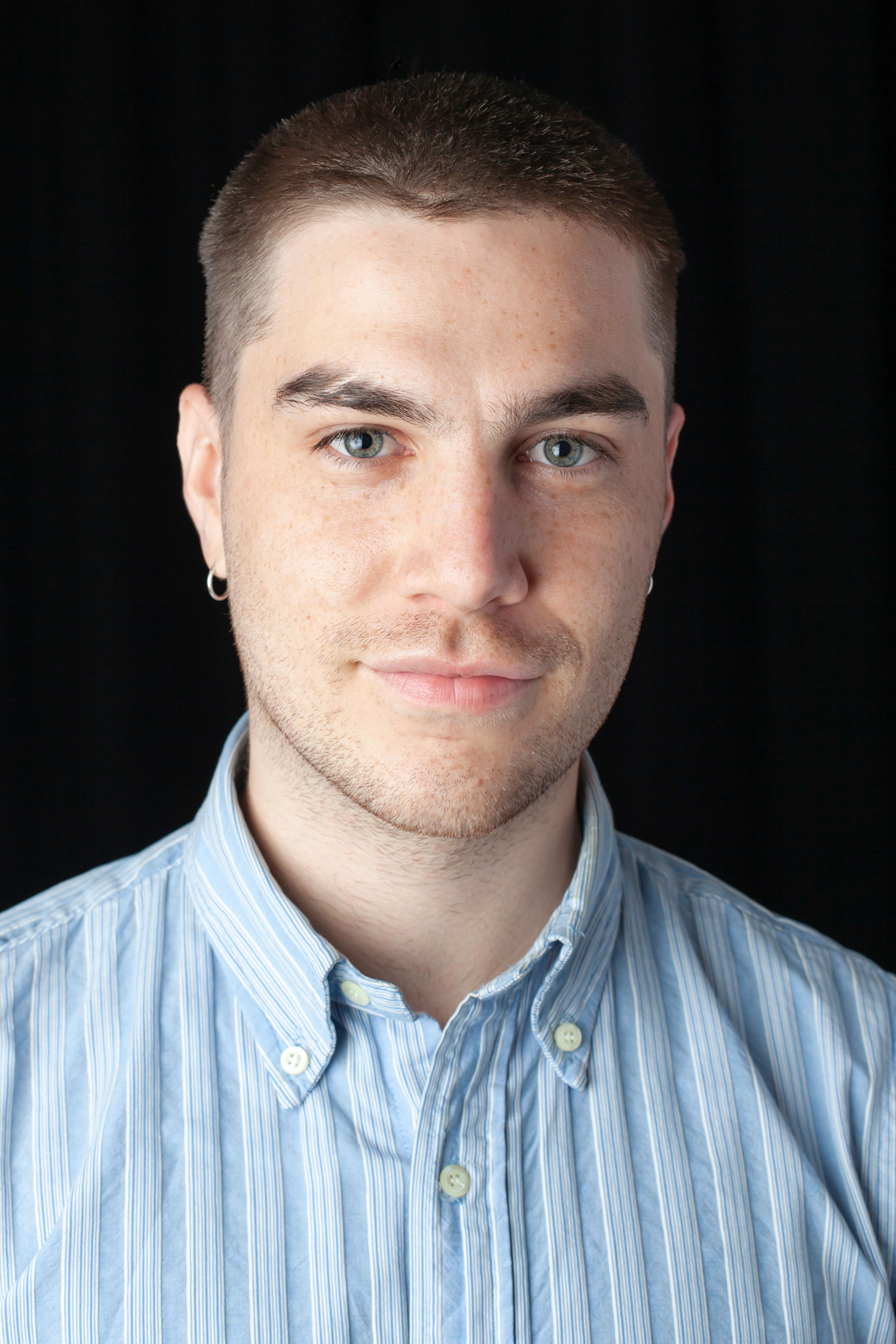 A portrait of a person with short hair, wearing a light blue striped button-up shirt, looking directly at the camera with a neutral expression