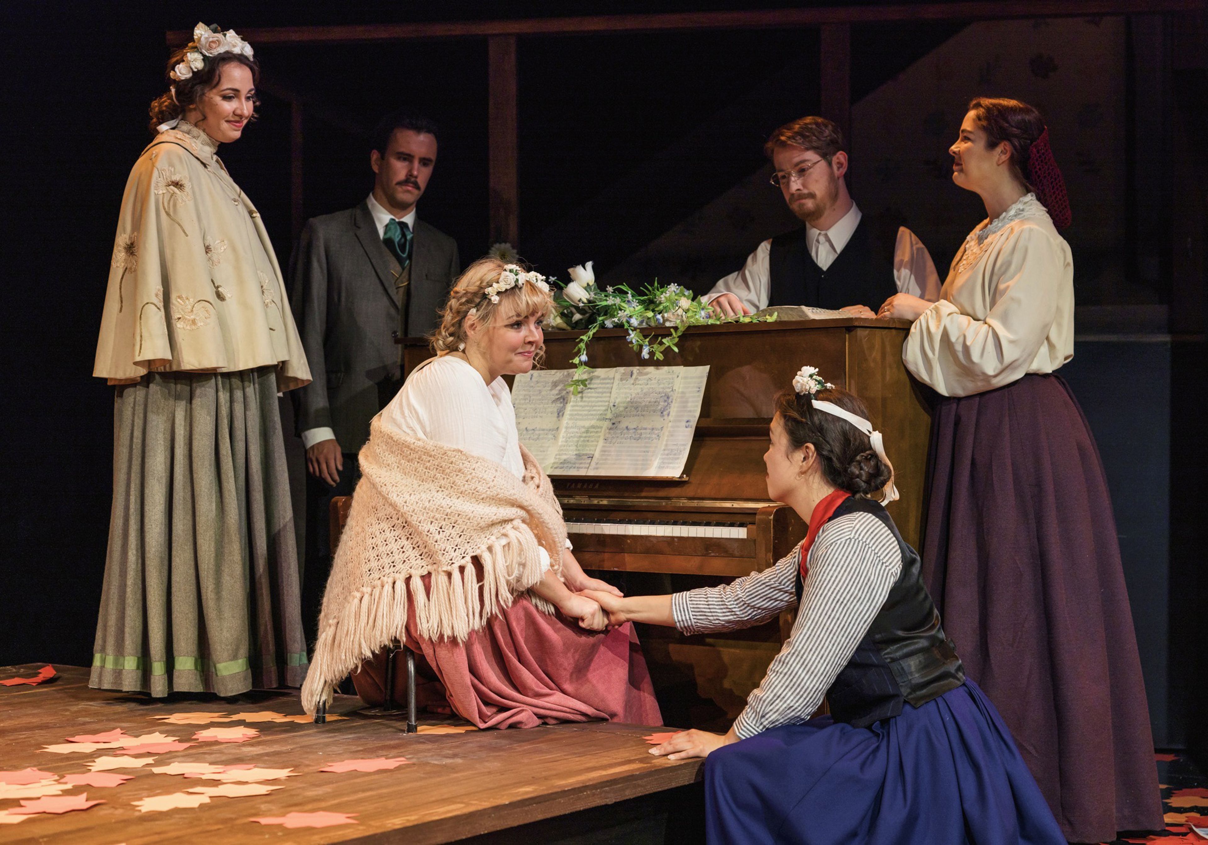 Six performers in period costumes gathered around a piano adorned with floral decorations. Two women sit in the foreground, holding hands in an emotional moment, while the others stand behind the piano observing