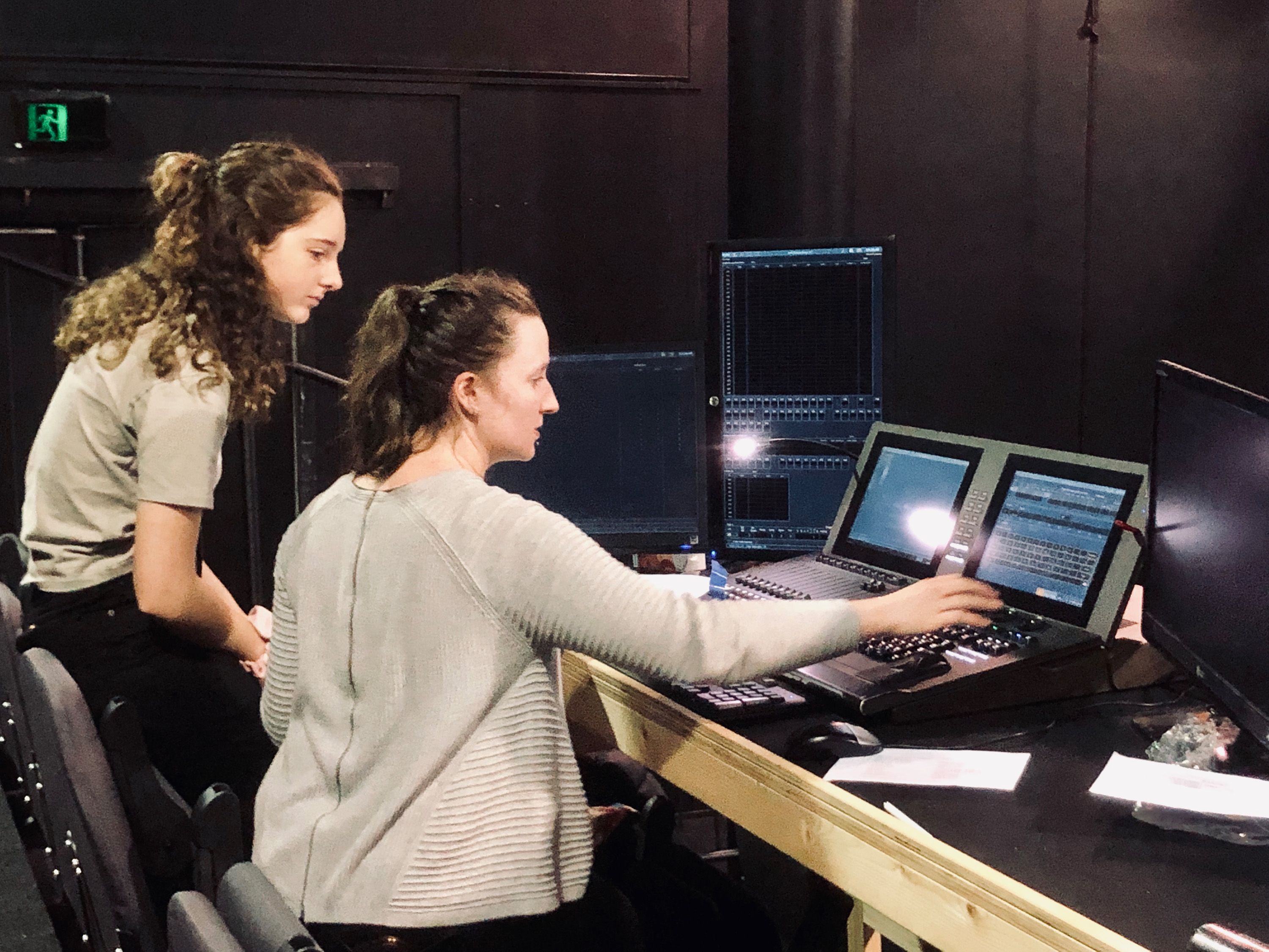 Two individuals working in at production desk on lighting console, one person is seated while the other stands nearby, observing.