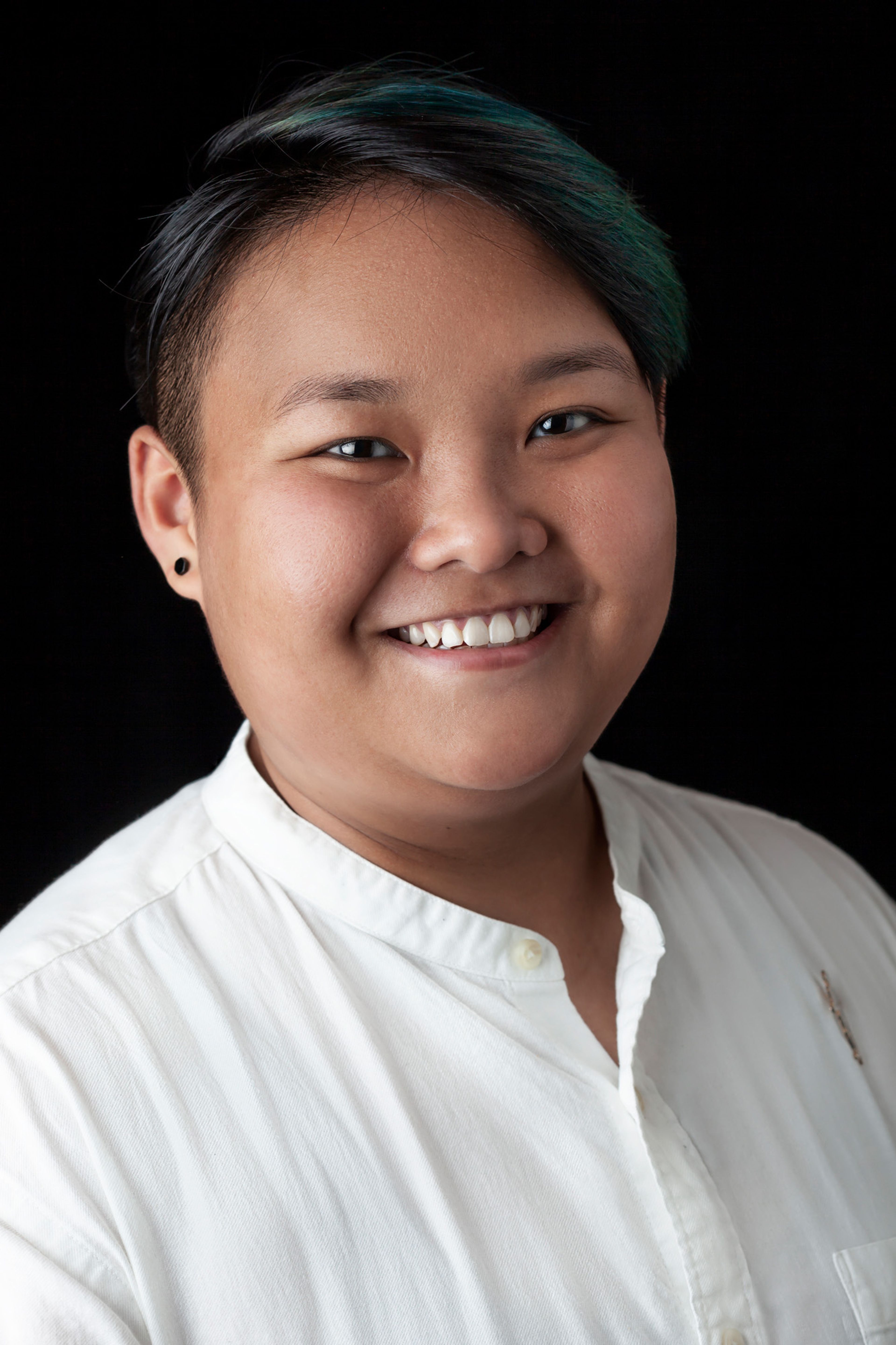 Portrait of a person with short, dark hair smiling. They are wearing a white button-up shirt and are photographed against a black background