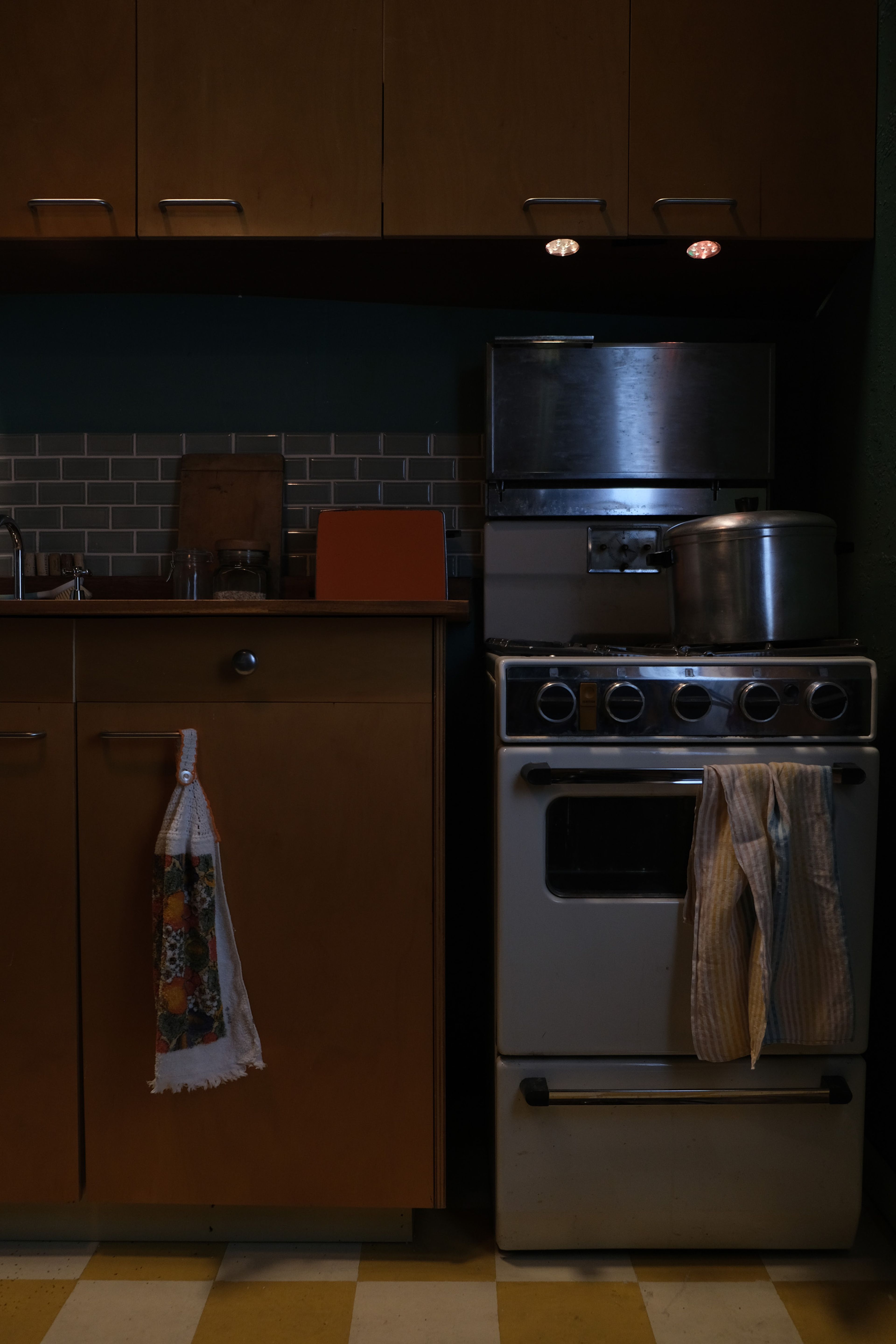 A dimly lit vintage kitchen scene featuring a wooden cabinet, tiled backsplash, and an old-fashioned stove with a large pot on top. A dish towel hangs from the stove, and a patterned towel is draped on a cabinet handle