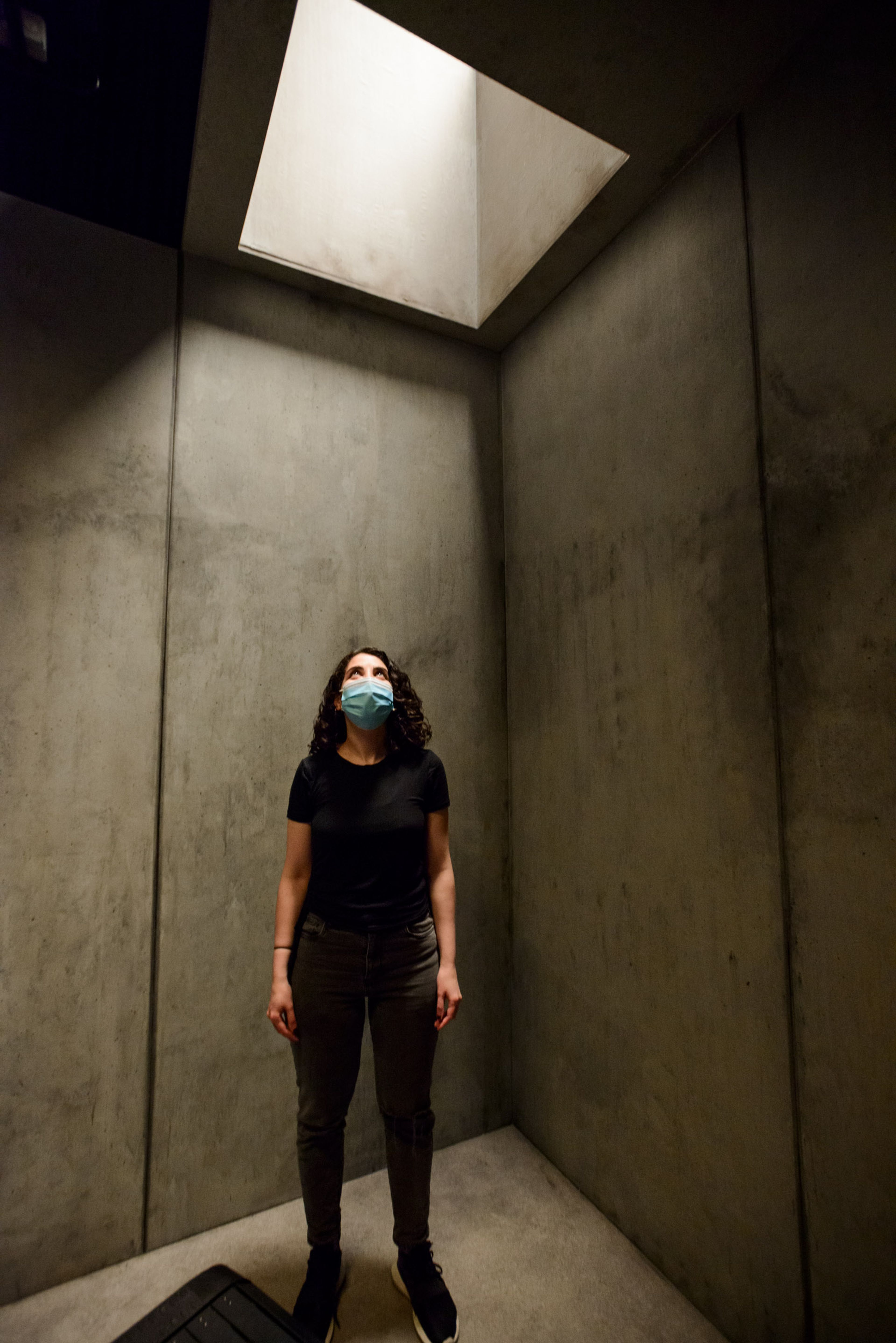 A person stands in a minimalist concrete room, illuminated by light streaming through a skylight above. They are wearing a face mask, black t-shirt, dark pants, and sneakers, gazing upward toward the light