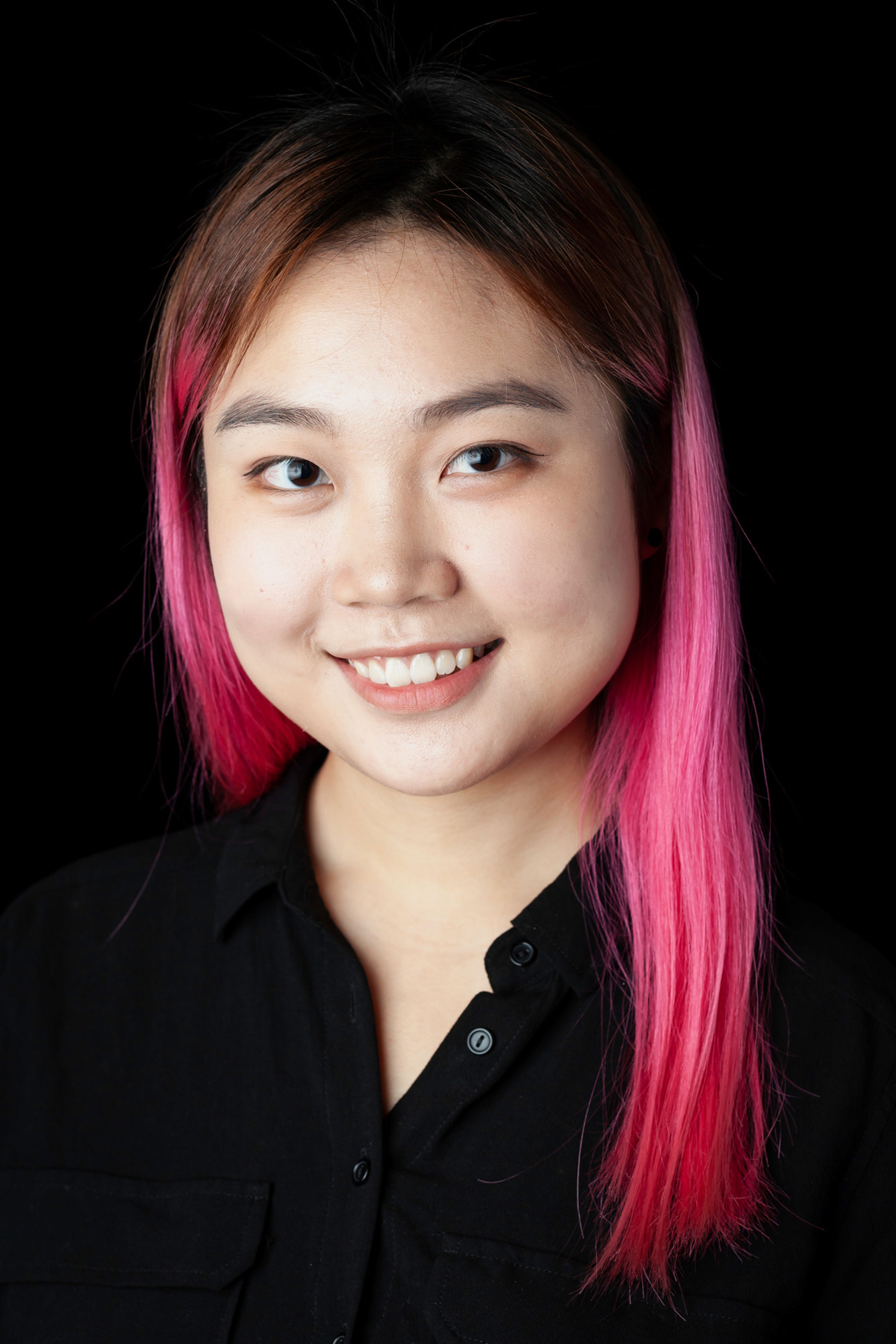 A portrait of a young woman with vibrant pink hair and a cheerful smile, wearing a black button-up shirt