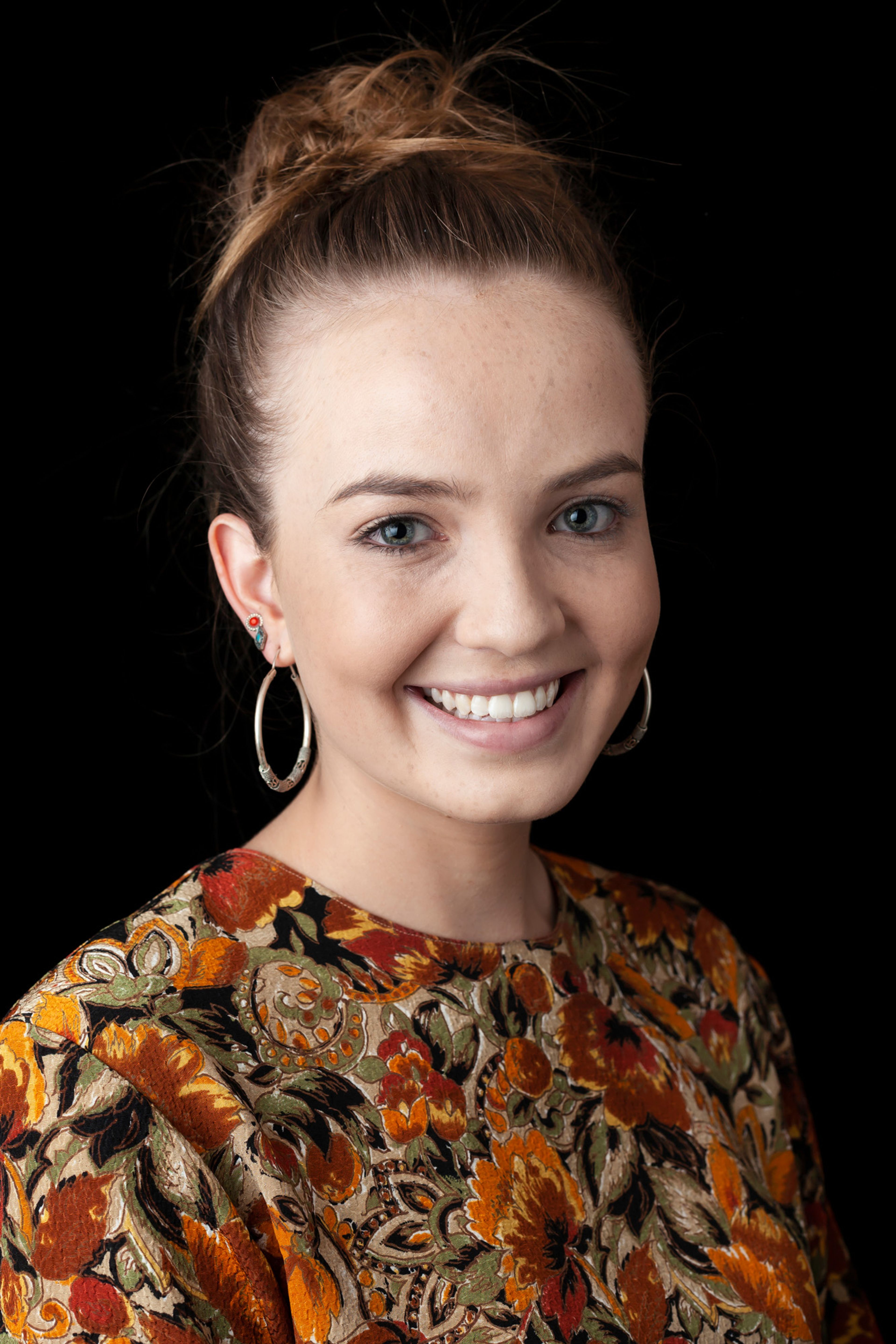 A portrait of a young woman with her hair in a bun, wearing large hoop earrings and a floral-patterned top with warm autumnal tones, smiling brightly