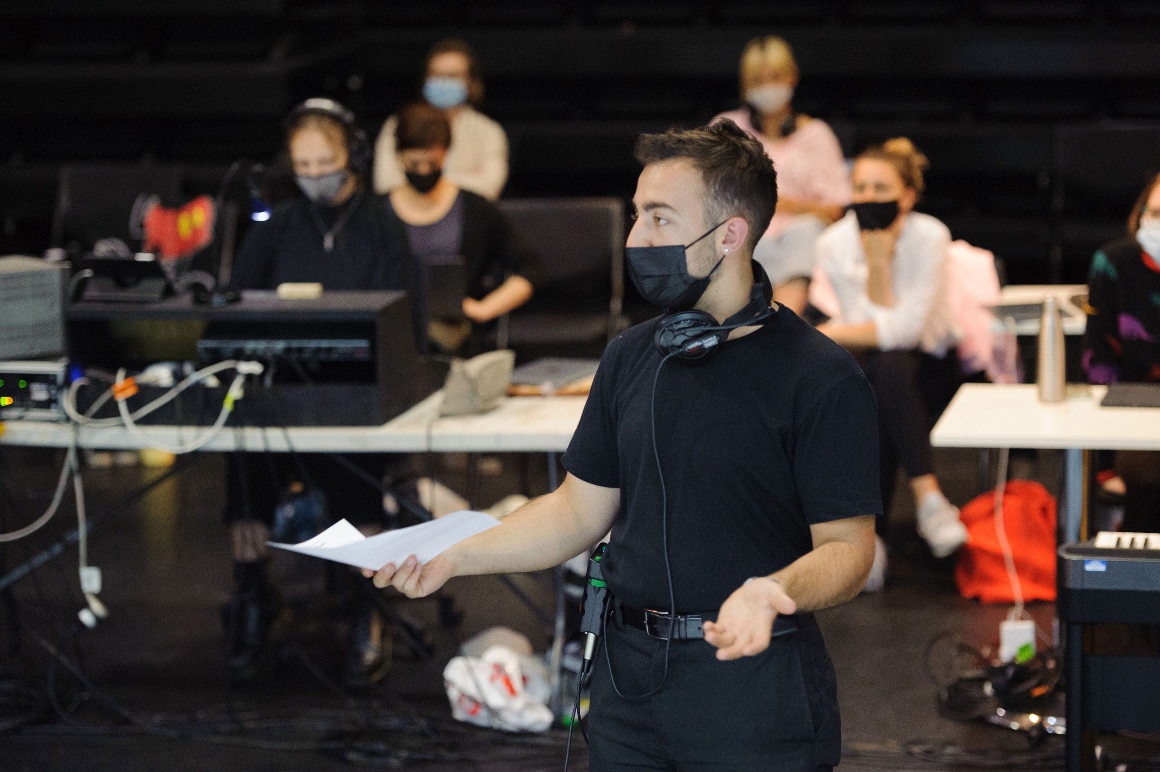 A person speaking and gesturing, with a production team working in the background