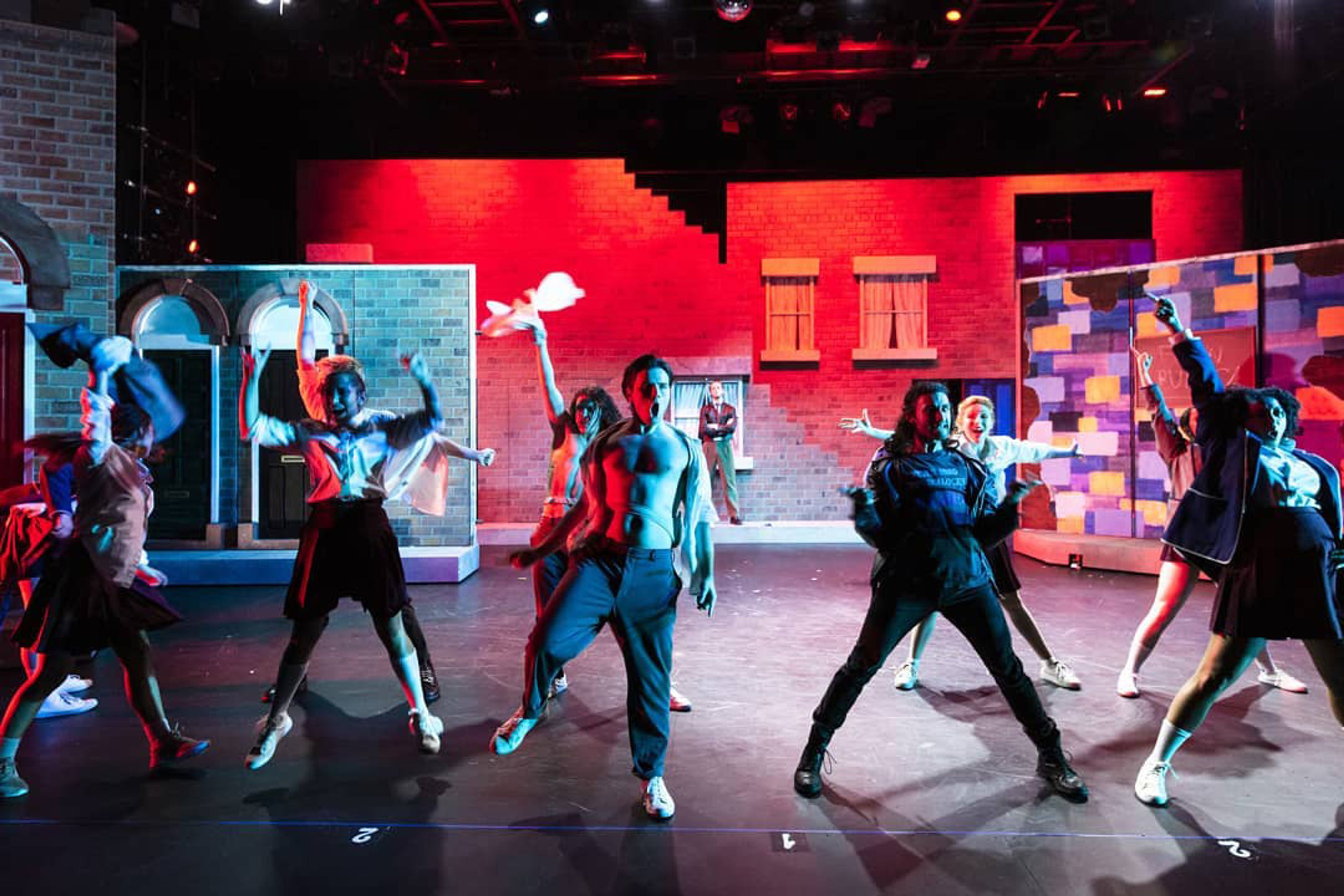 A dynamic stage performance with performers in expressive poses, vibrant red and blue lighting, and a backdrop of brick facades and colorful geometric patterns.