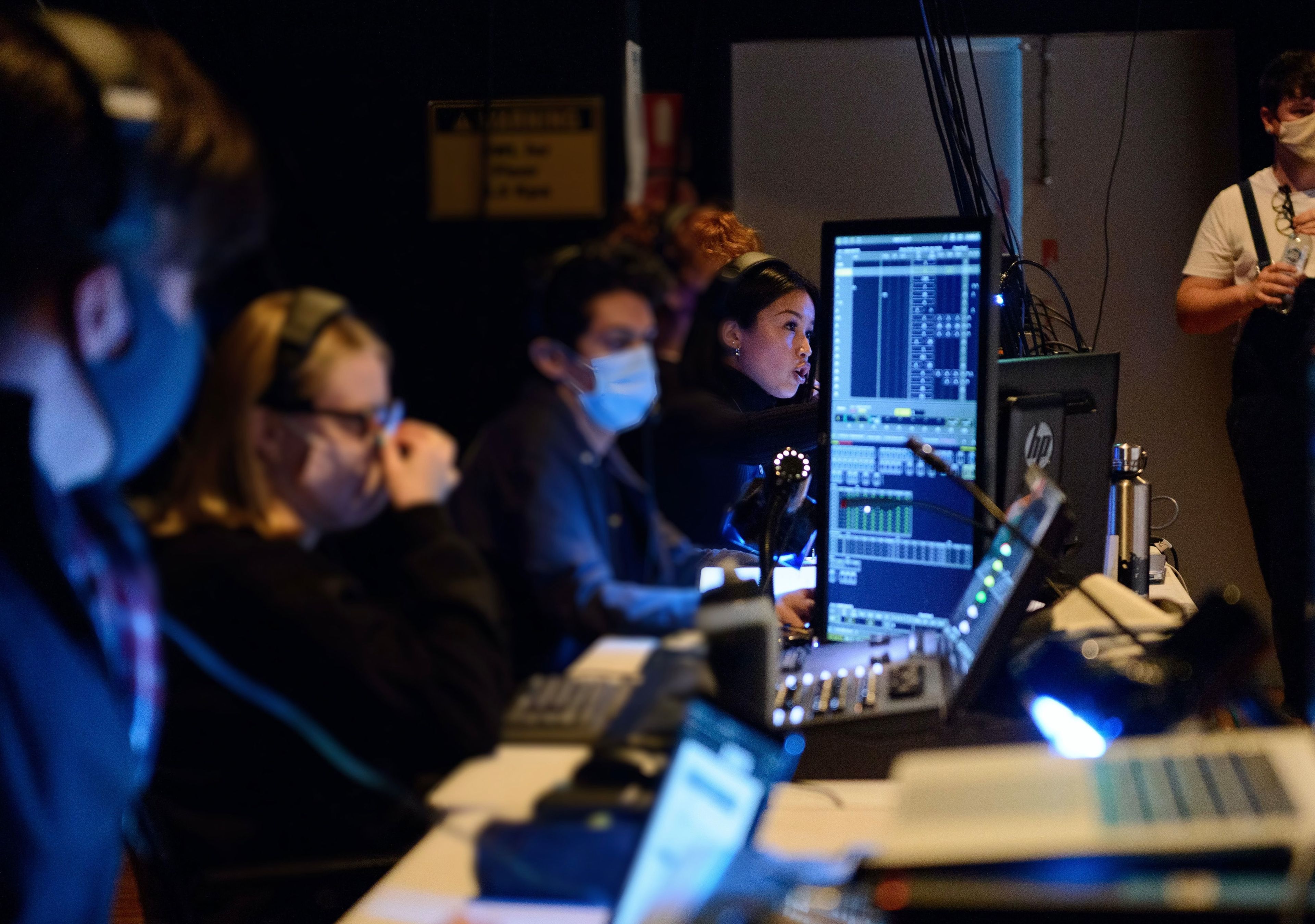 A backstage production team, masked and focused, operating sound and lighting equipment in a dimly lit theatre